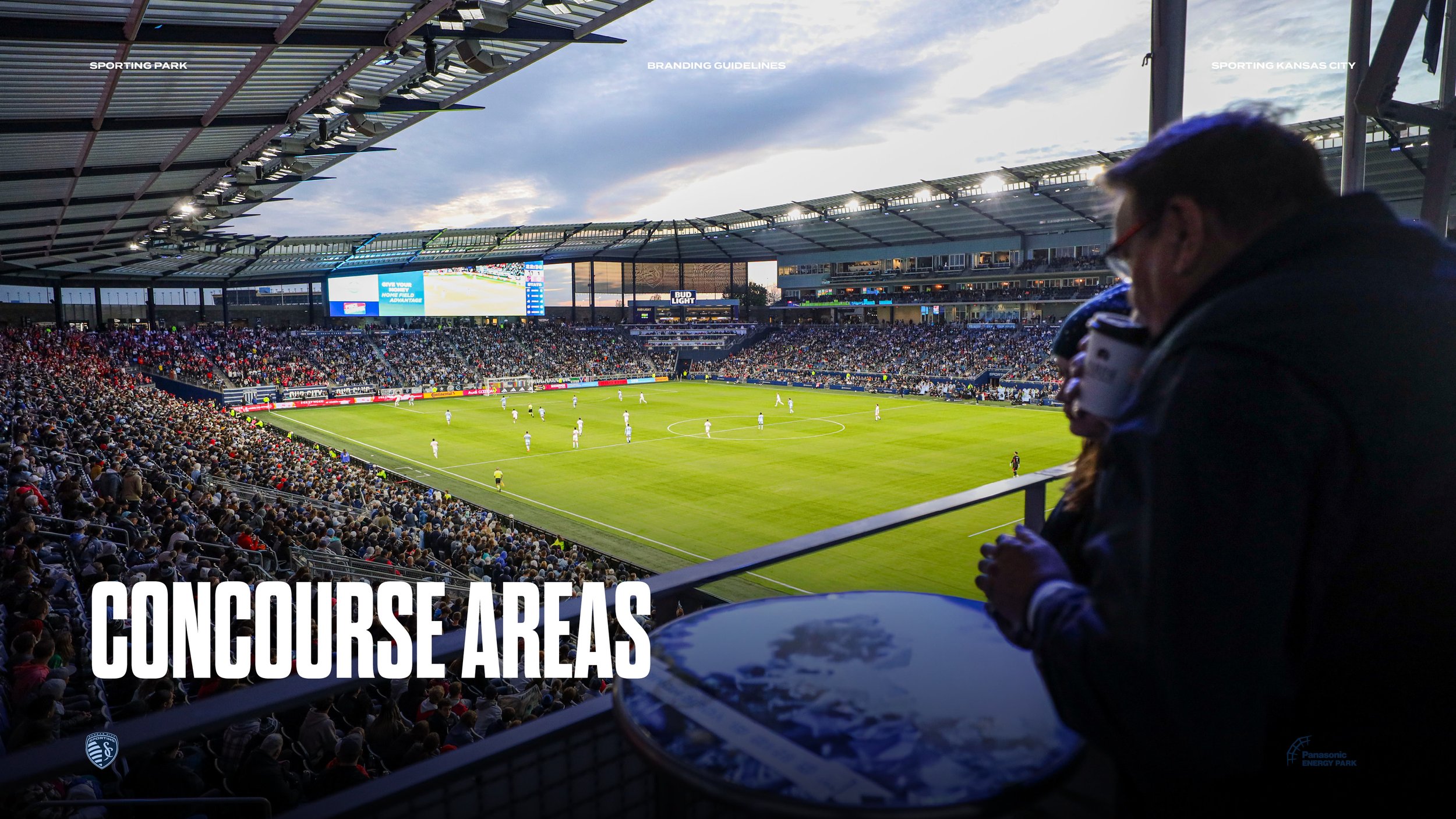 A packed soccer stadium during a game at Panasonic Energy Park in Kansas City, with players on the field and a large crowd in the stands. A person is sitting in the foreground, possibly a commentator or announcer, holding notes or a microphone, with 