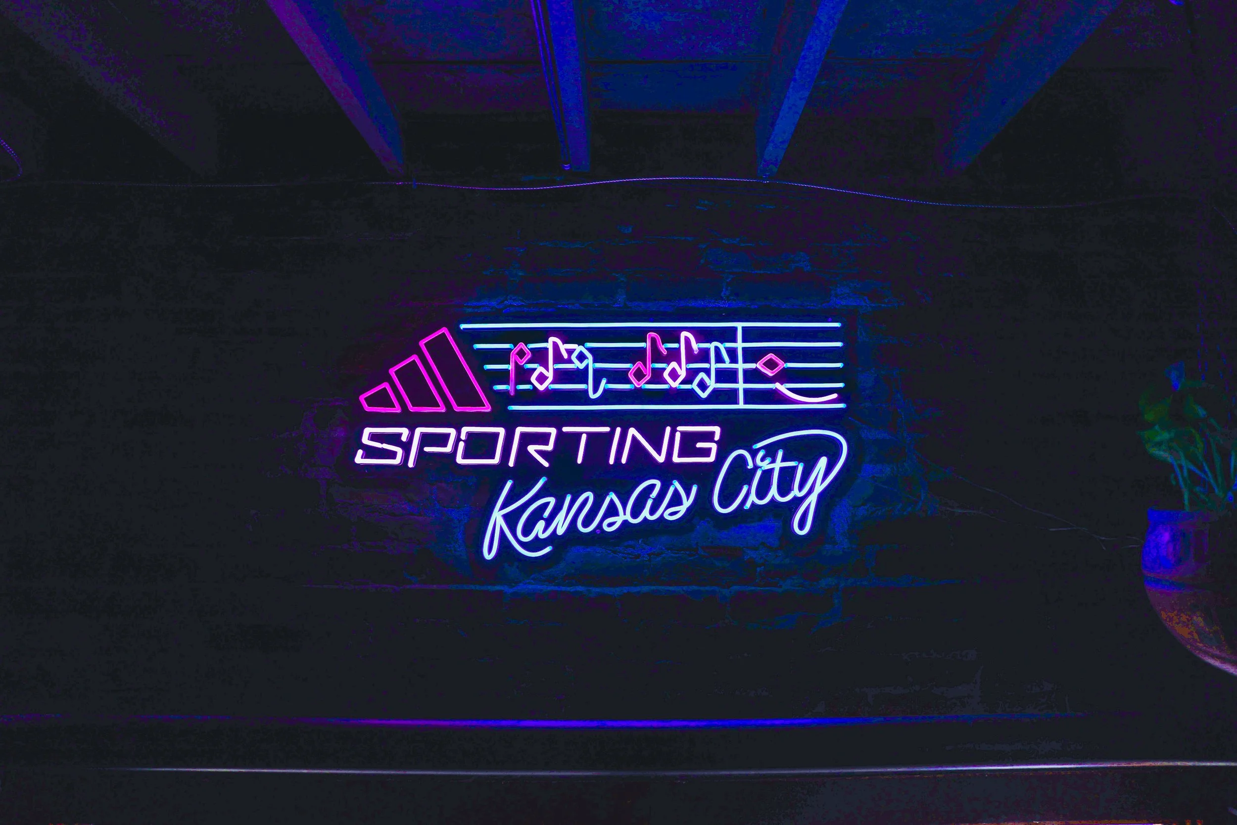 Neon sign with musical notes, the Adidas logo, and the words 'Sporting Kansas City' in purple and blue neon lights on a dark brick wall.