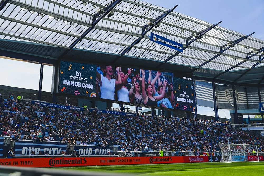 A large crowd of people in a stadium, watching a cheerleading or dance performance on a big LED screen. The screen displays young girls cheering with their hands raised. The stadium has a sign that reads "Childrens Mercy Park."