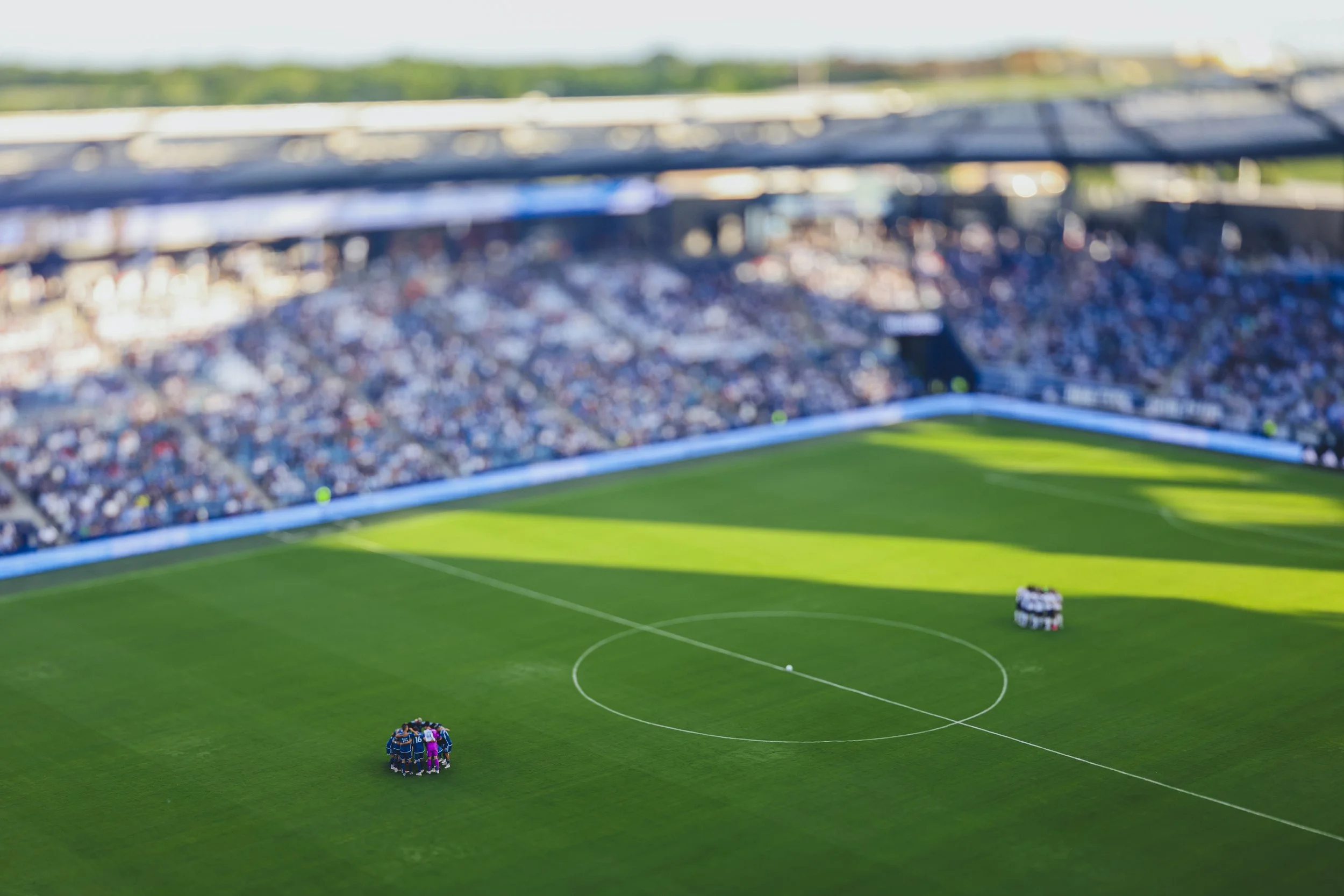 A soccer stadium with spectators, two teams of players huddled together on the field, with bright sunlight casting shadows.