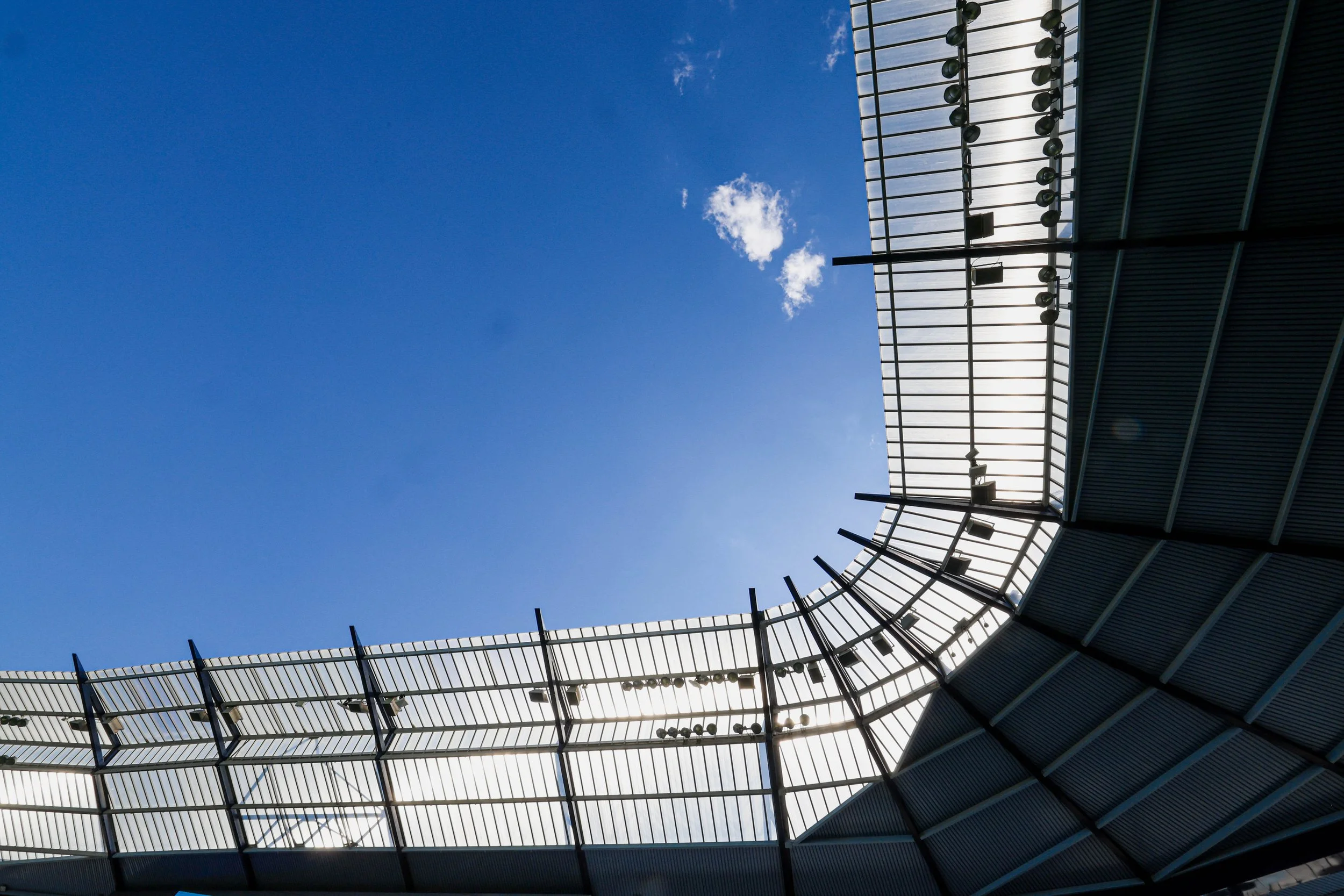 Looking up at a curved stadium roof with metal framework and lights, against a bright blue sky with a few clouds.