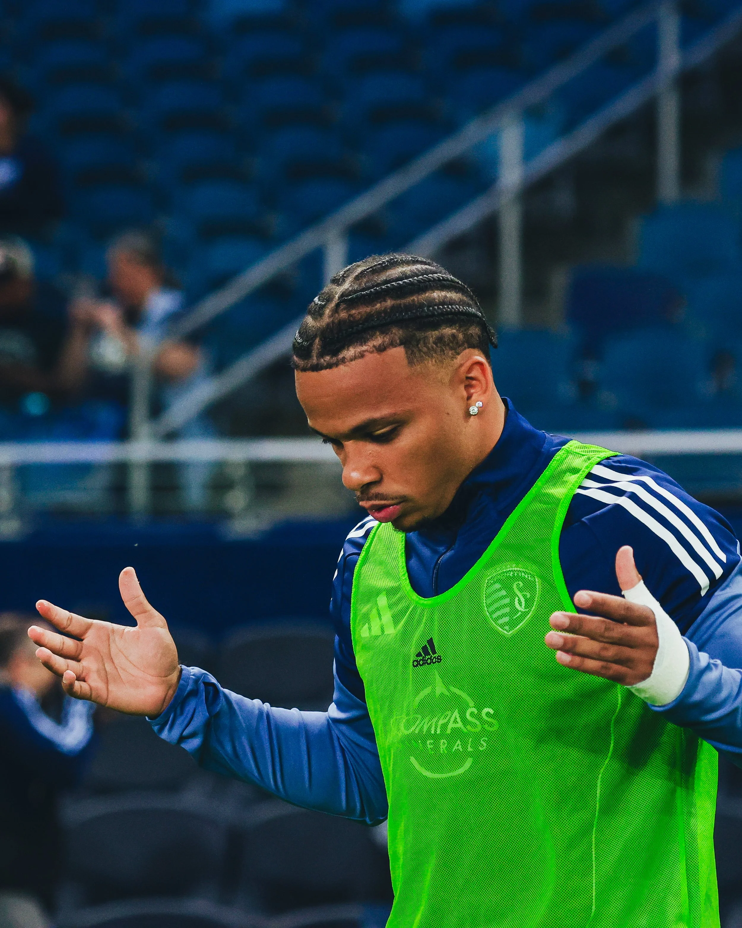 A soccer player with braided hair and earrings wearing a blue jersey and a green training bib, practicing on a field.