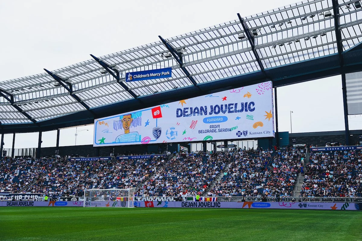 View of a soccer stadium filled with spectators, a large digital screen displaying a graphic with the name 'Dejan Jovelic', indicating he is a forward from Belgrade, Serbia, with a drawing of a person and colorful decorations, under the sign 'Children's Mercy Park'.