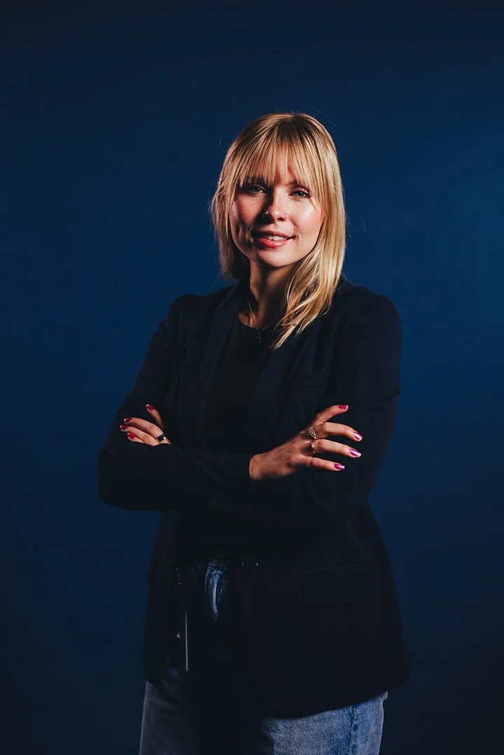 A woman with blonde hair and bangs wearing a black blazer and jeans, crossing her arms, standing against a dark blue background.