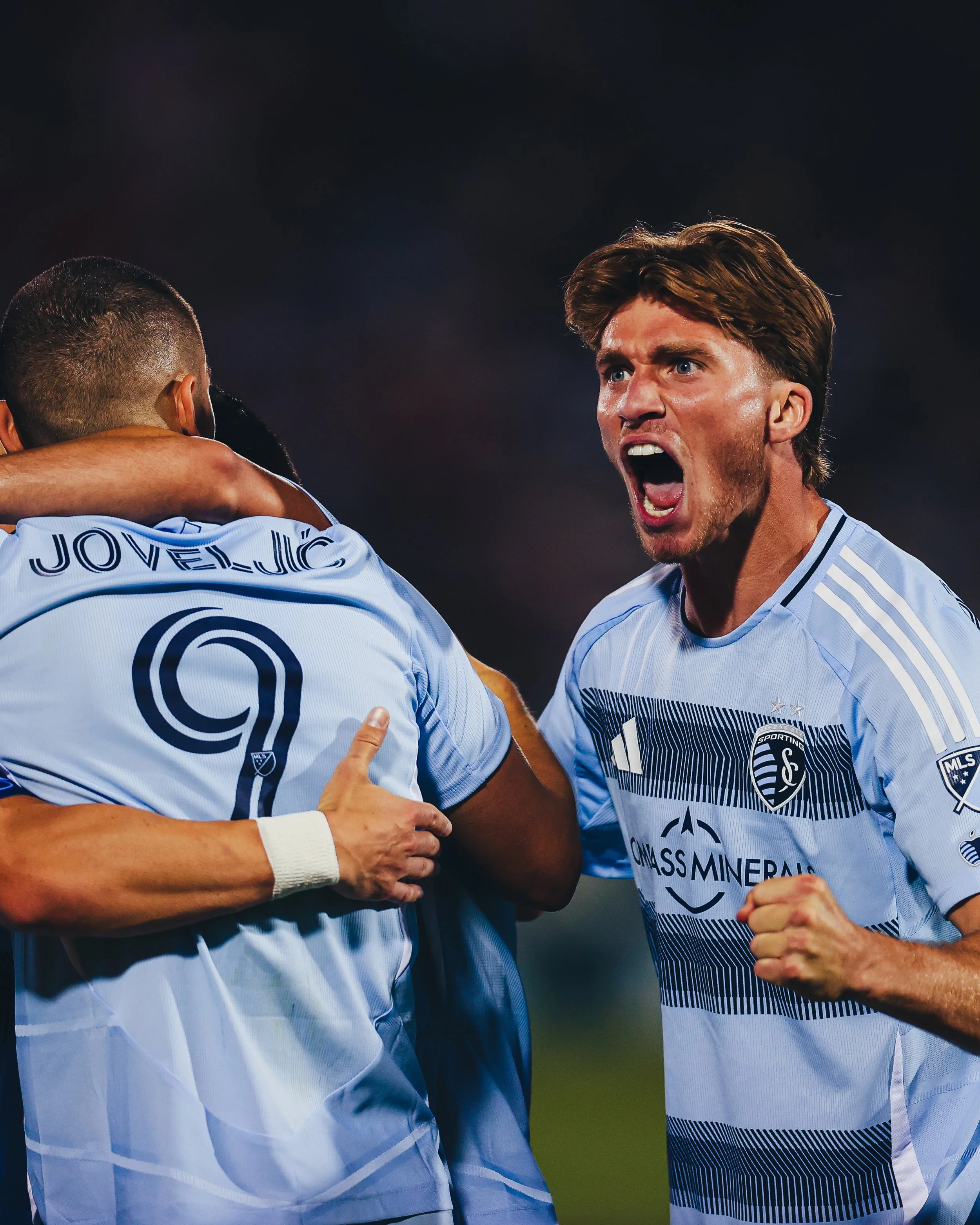 Soccer players celebrating a goal, with one player shouting and another with his arm around a teammate.