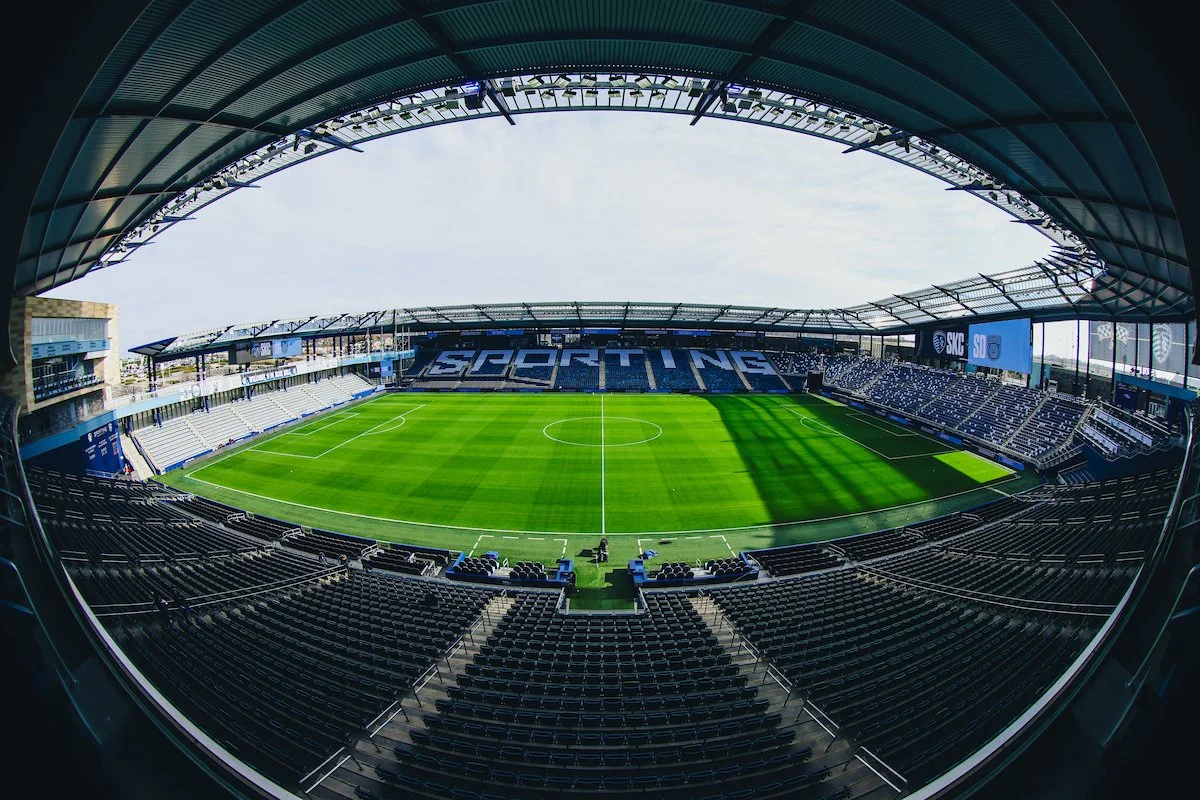Empty soccer stadium with green field and black seats, partially covered roof, and large screen displays.