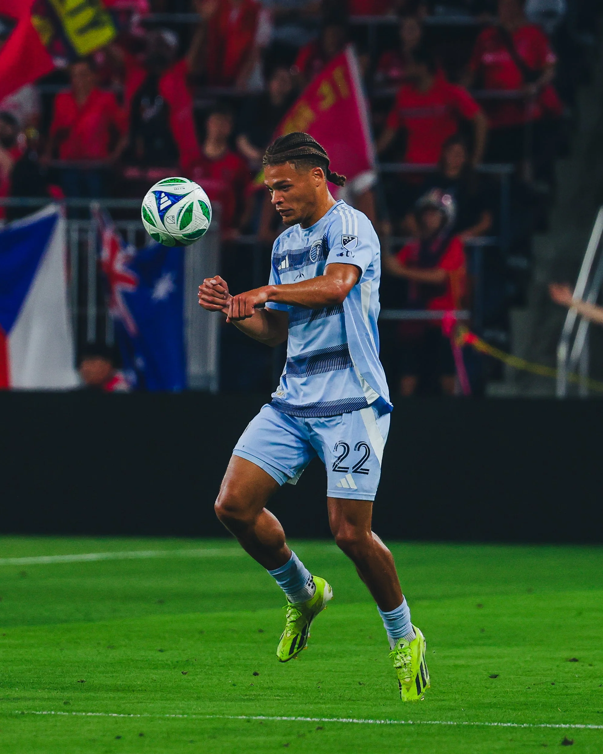 A soccer player in a light blue uniform with the number 22 is on the field, watching a soccer ball in midair during a game.
