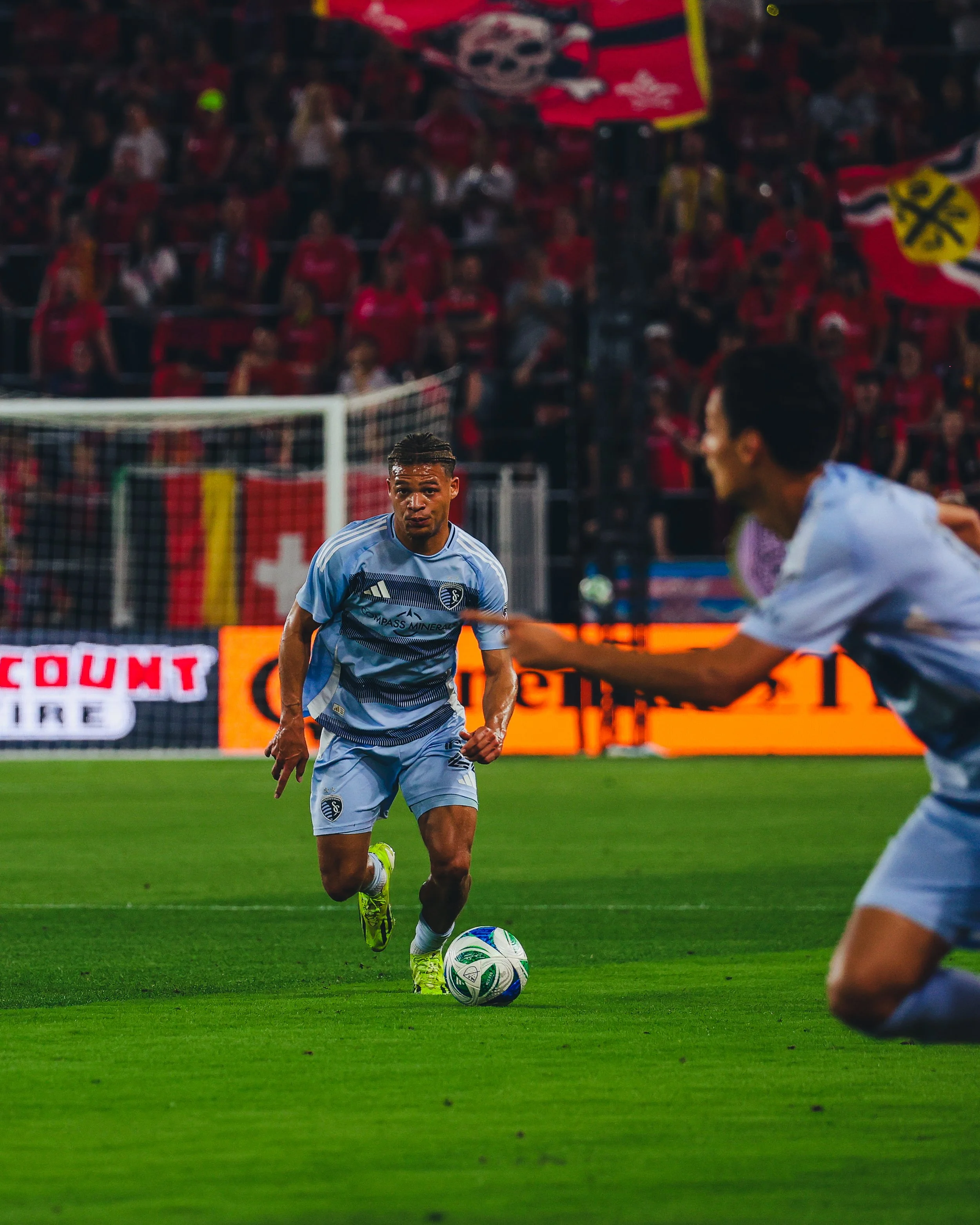 A soccer player in a light blue uniform dribbling a ball on the field during a match, with an opponent rushing towards him and a crowd in the stands cheering in the background.