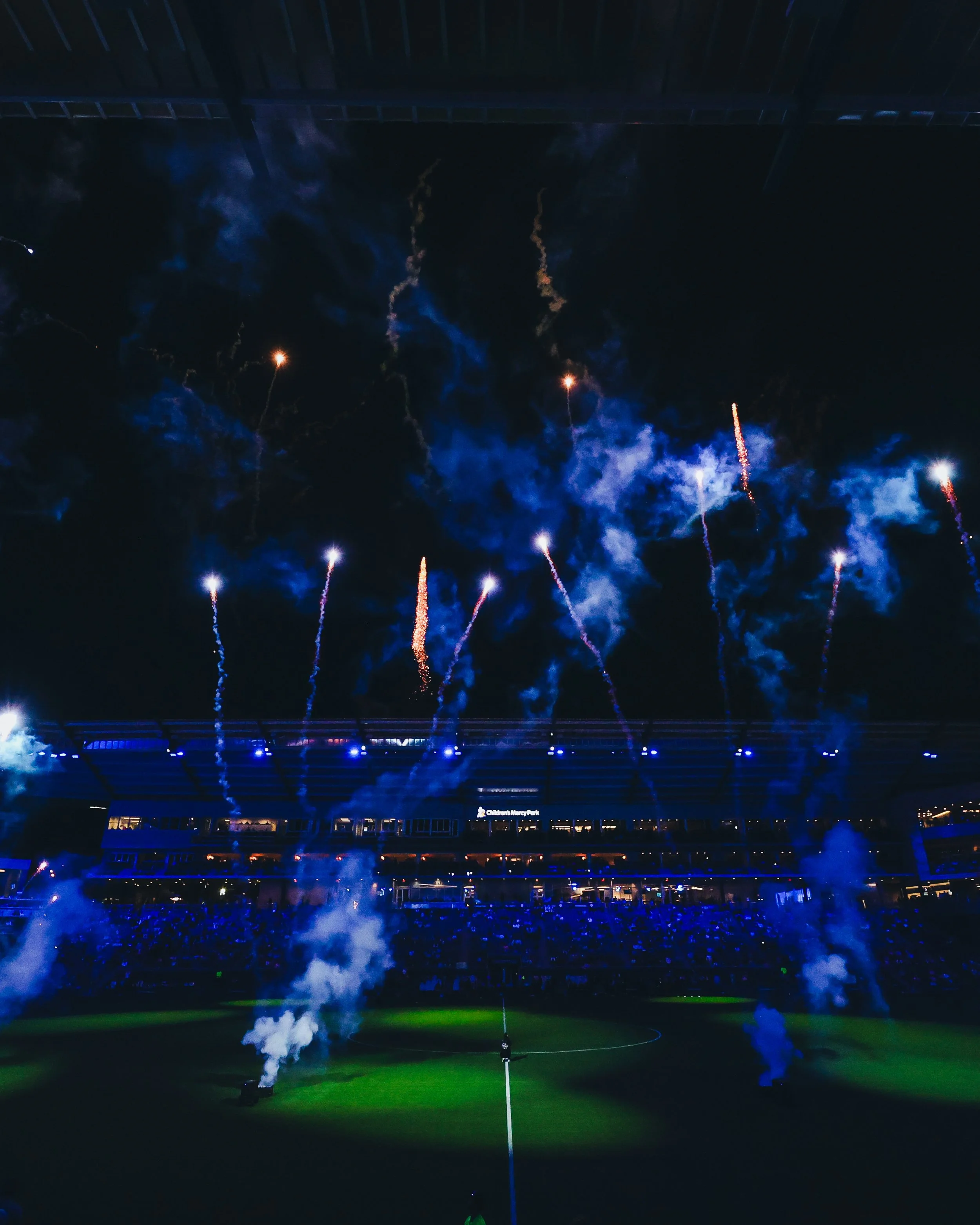 Fireworks display over a stadium at night with smoke and bright lights.