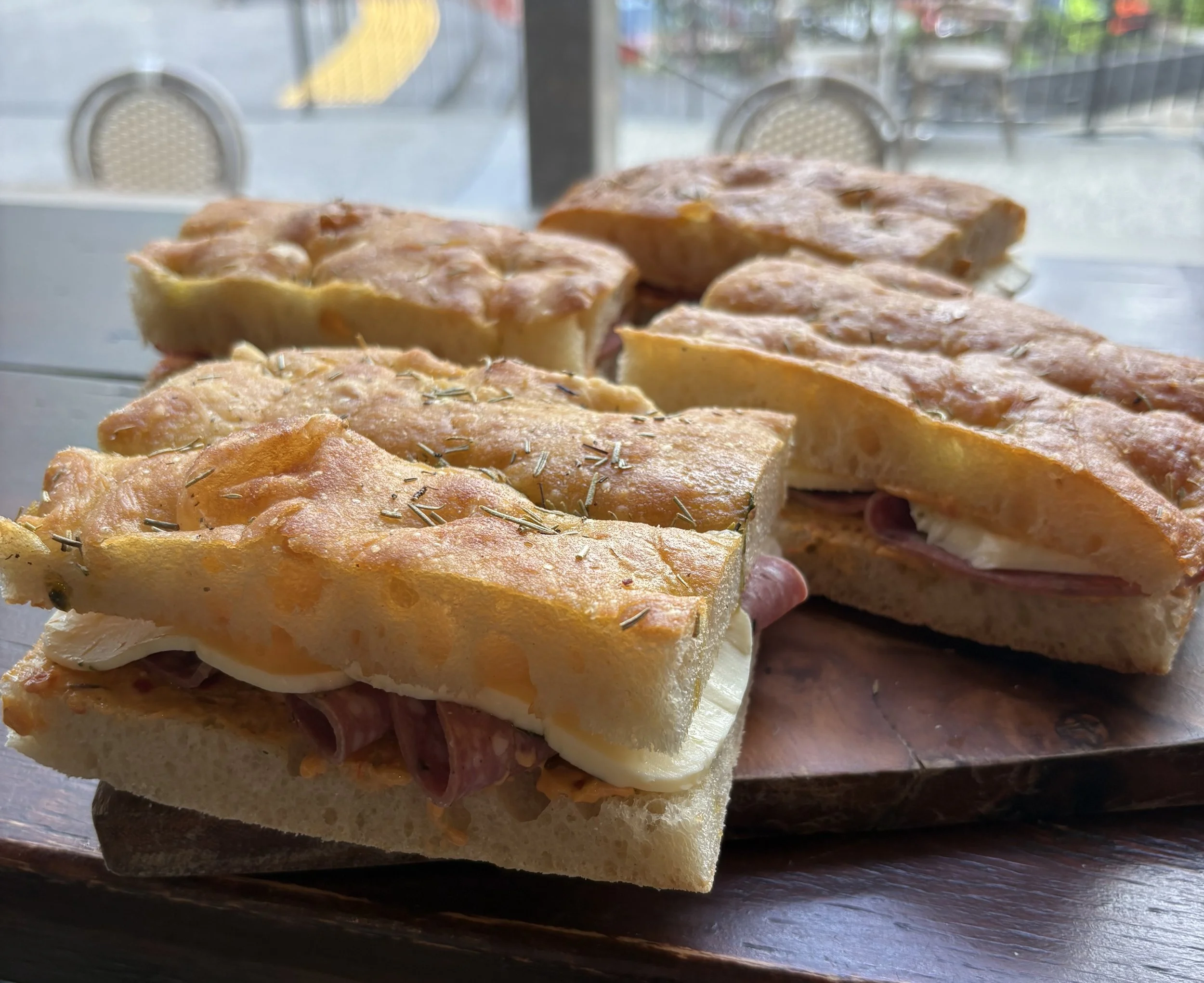 A close-up of a sandwich with melted cheese, ham, and herbs on a wooden board, set in front of a window with a view of chairs and outdoor scene.