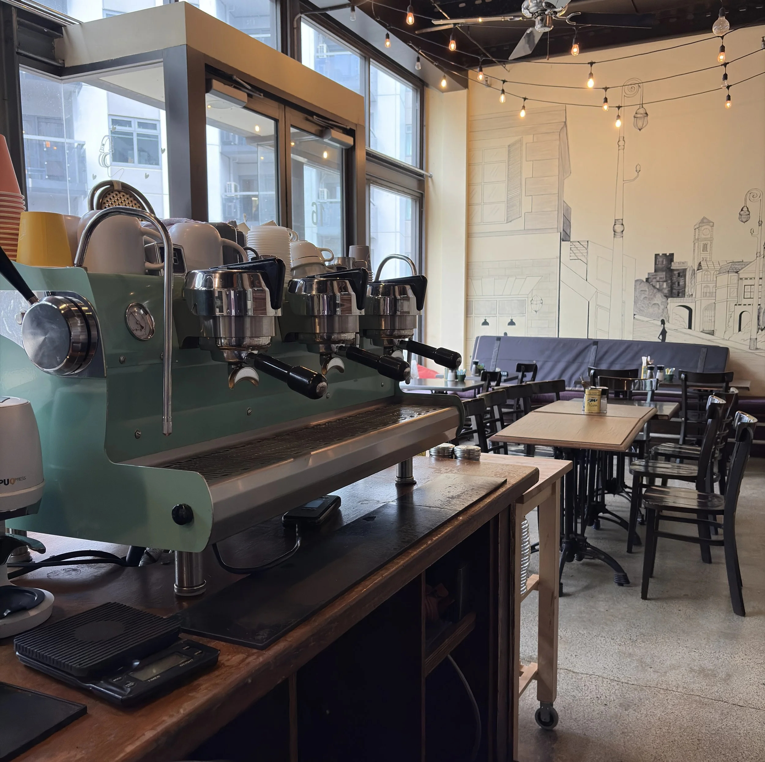 A coffee shop interior with a wooden counter, espresso machine, stacks of cups, and coffee accessories. Large windows letting in natural light, wall art depicting city buildings, and string lights hanging from the ceiling.