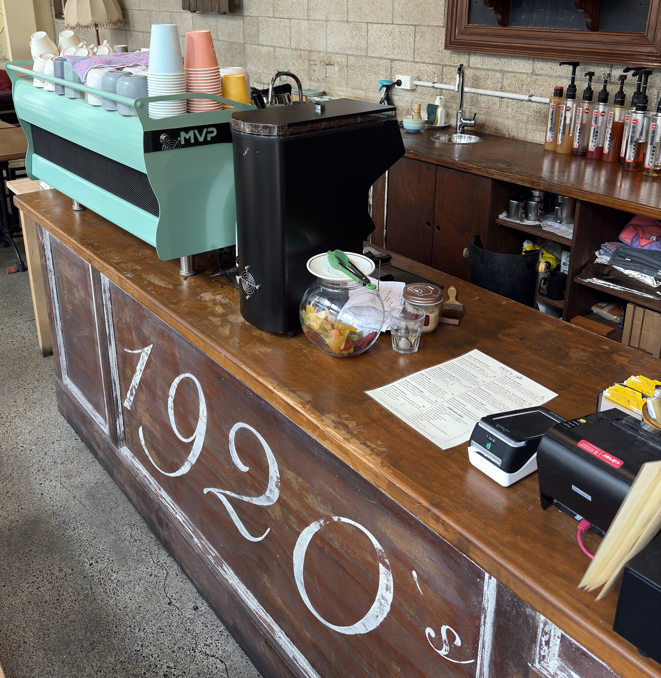 Counter with colorful cups, a coffee machine, and various bottles of flavored syrup, with a coffee menu and utensils.