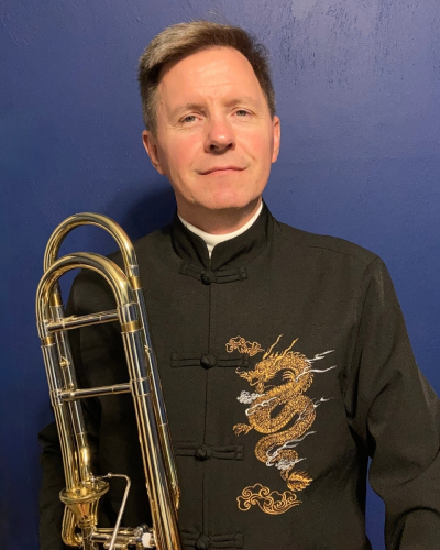 A man in a black traditional Chinese jacket with gold dragon embroidery, holding a trombone, standing against a blue wall.