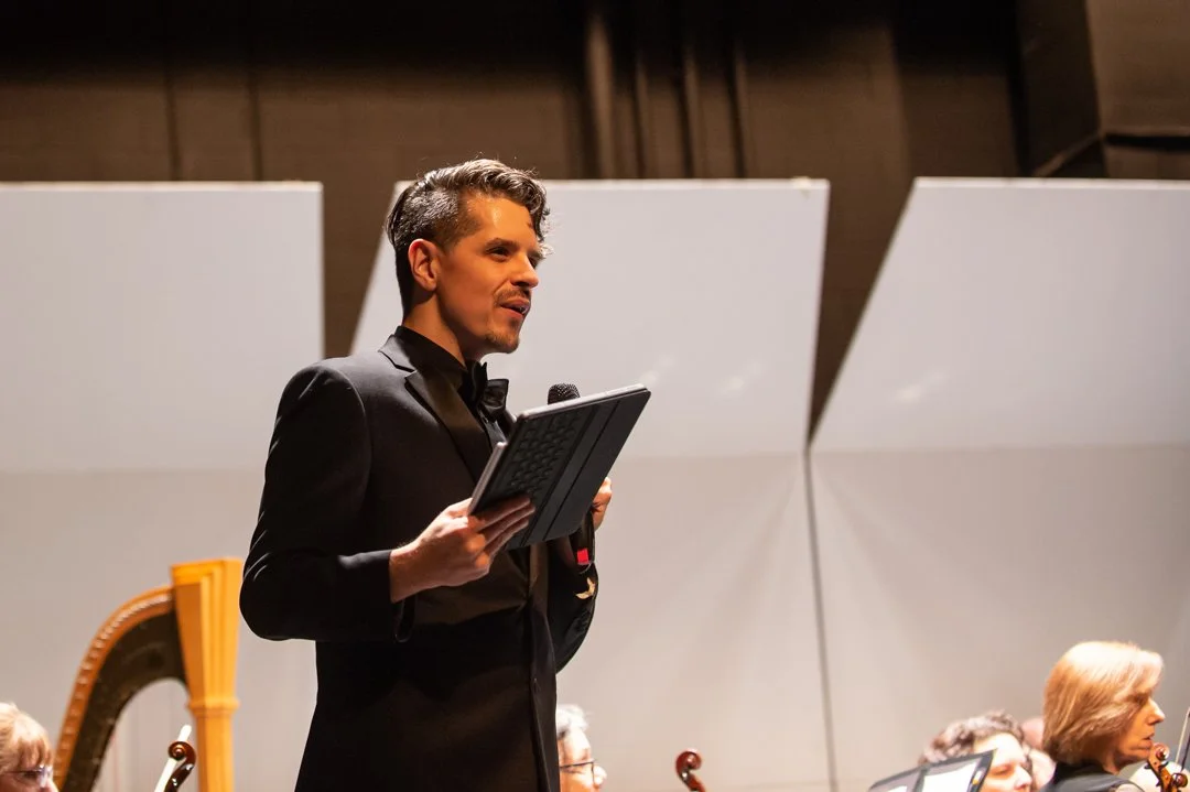 A male conductor in a black tuxedo holding a baton and a clipboard, leading an orchestra on stage.