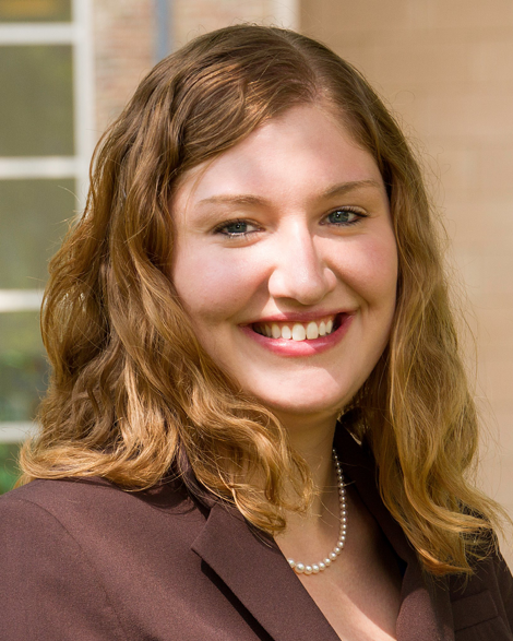 A smiling woman with shoulder-length wavy light brown hair, wearing a dark blazer and pearl necklace, outdoors with a building and window in the background.