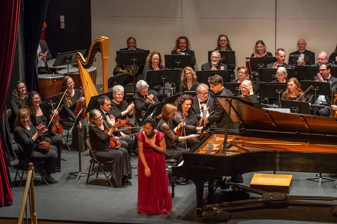 An orchestra on stage with a conductor, pianist, and a soloist in a red dress, applauding, with many musicians holding string instruments and a harp, in a concert hall.