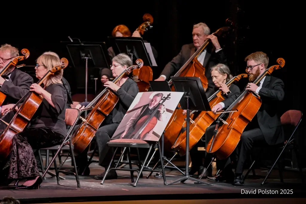 An orchestra performing on stage featuring string musicians playing violins, cellos, and double basses, with music stands and a photograph of a cellist on a chair in front.