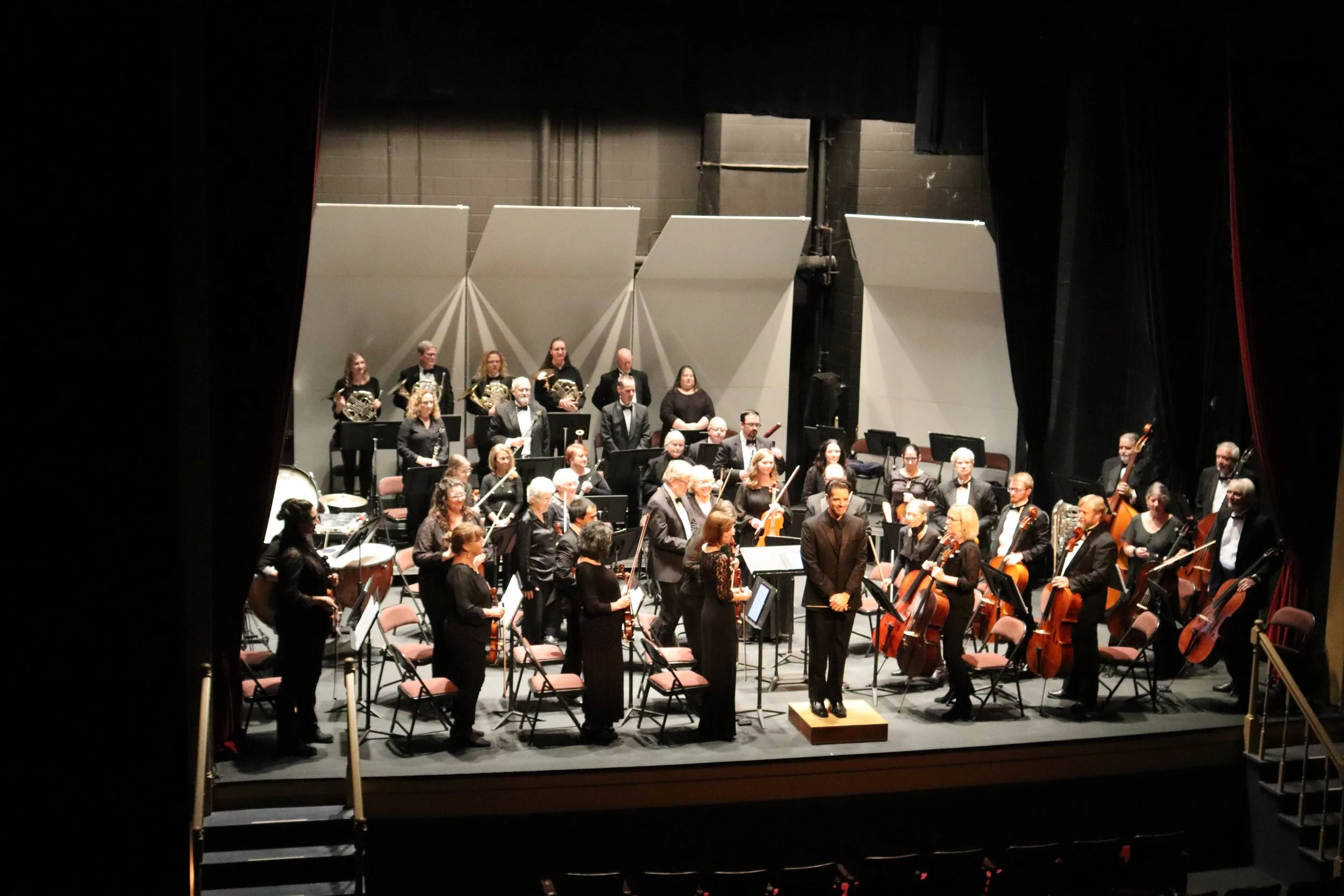 Orchestra members on stage preparing to perform, with some standing and others seated with musical instruments, view from behind the stage curtain.