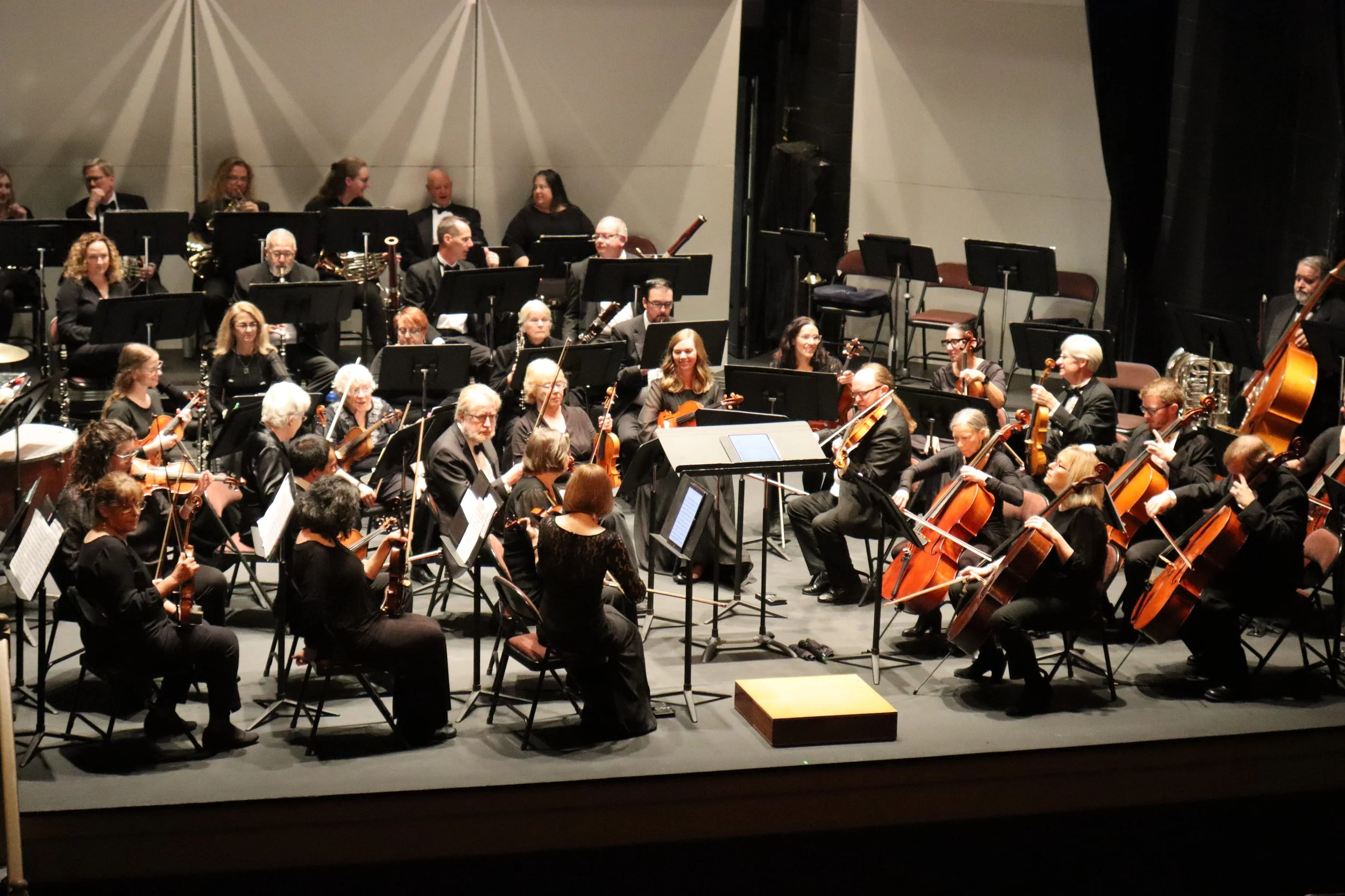An orchestra performing on stage with musicians playing various instruments including violins, cellos, and wind instruments, seated in black formal attire, with stage lights overhead illuminating the scene.
