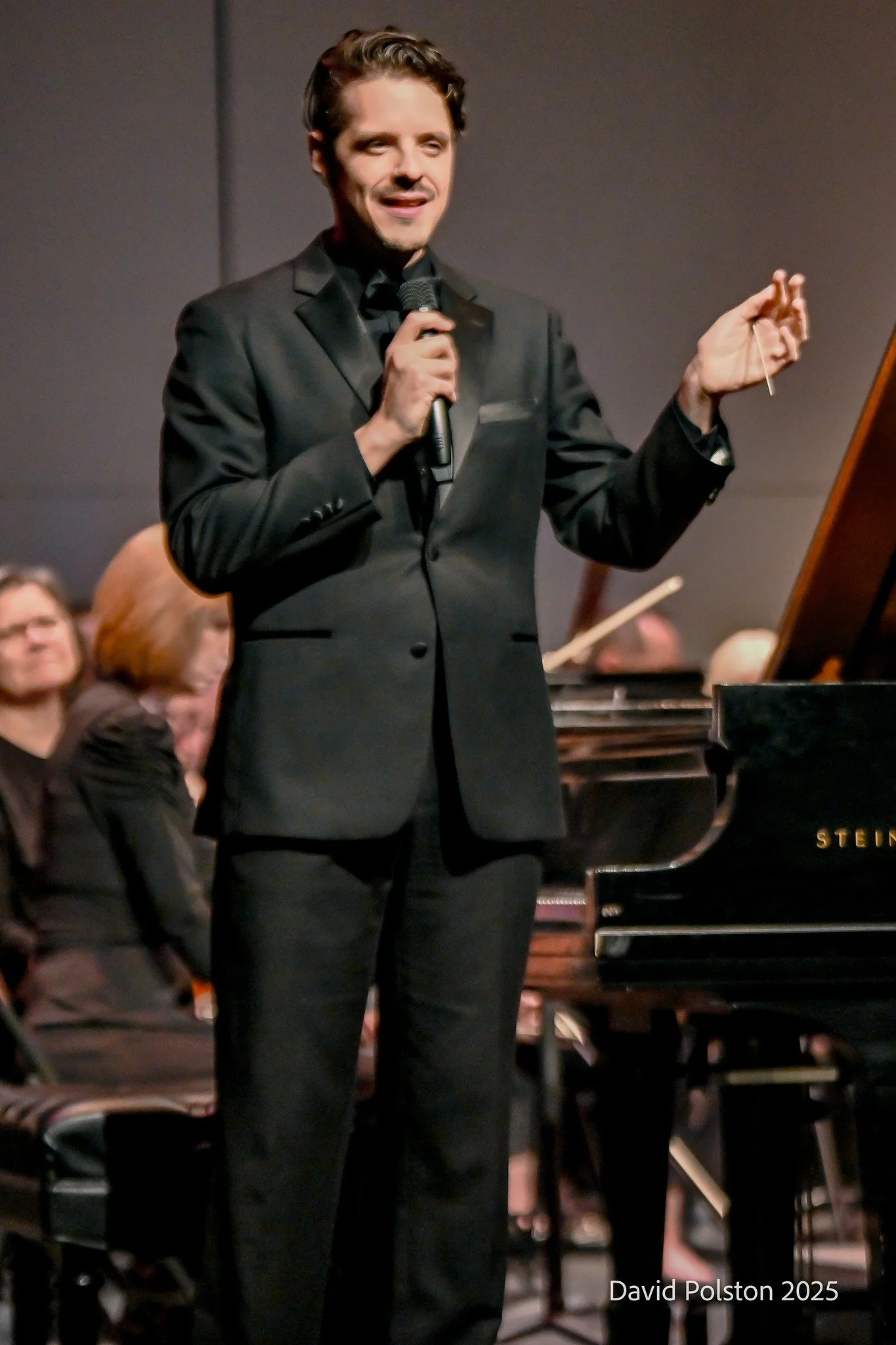 A man in a black suit holding a microphone and speaking during a musical performance. People are seated in the background with a grand piano visible. The photo is credited to David Polston 2025.