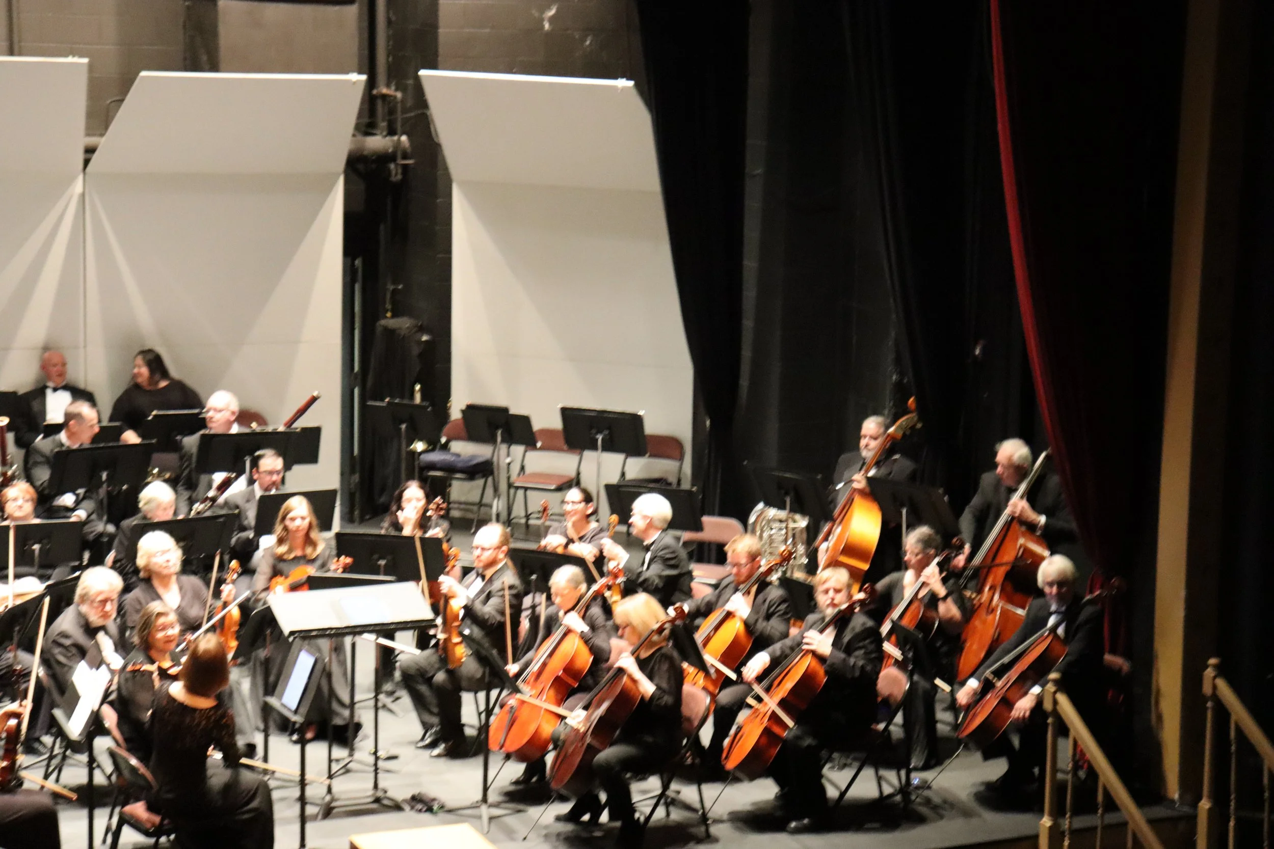 An orchestra performing on stage with musicians playing string instruments such as violins and cellos. The stage is set with black curtains and white backdrop panels.