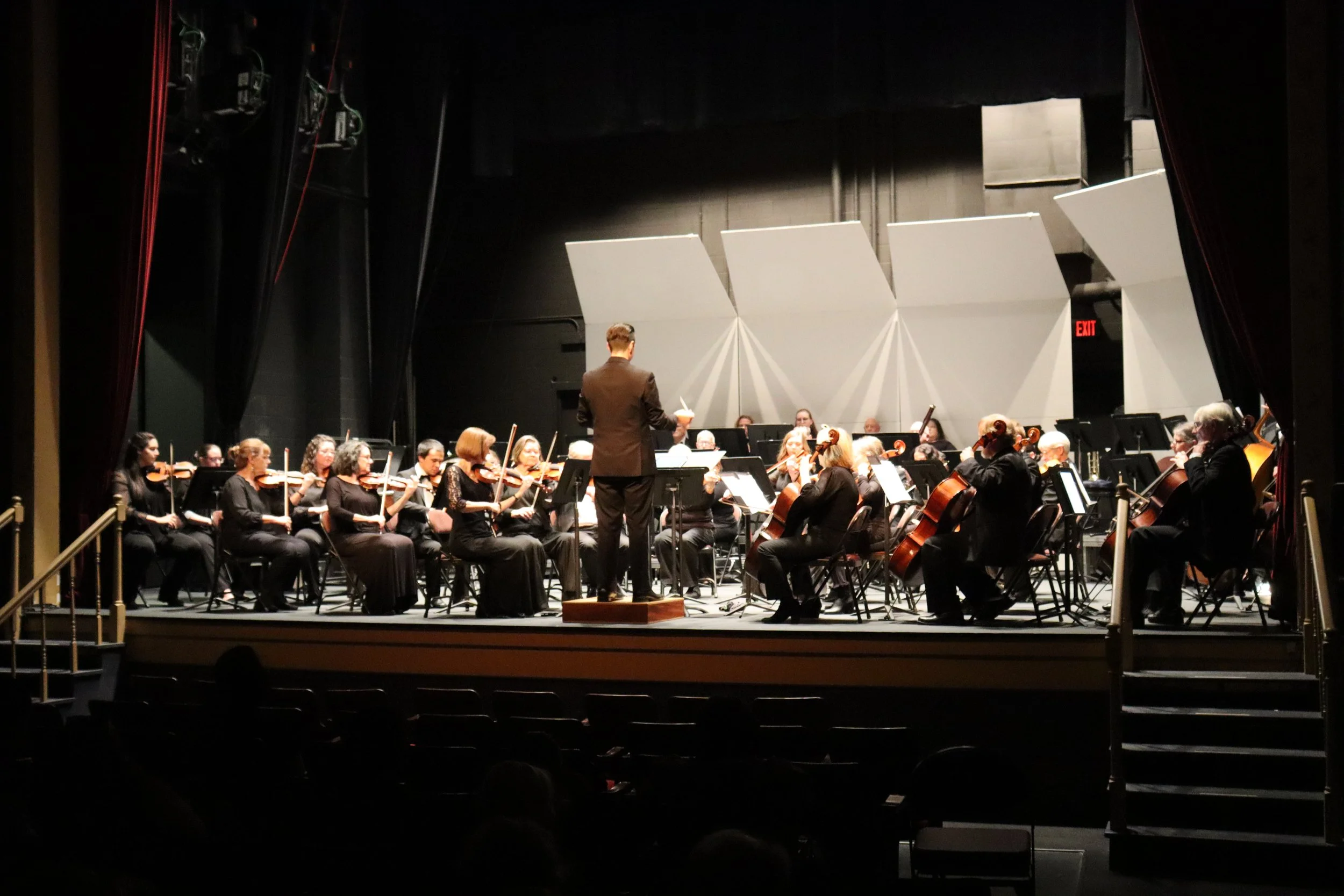 An orchestra performing on stage with a conductor leading them, in a theater with black walls and a partly open red curtain.