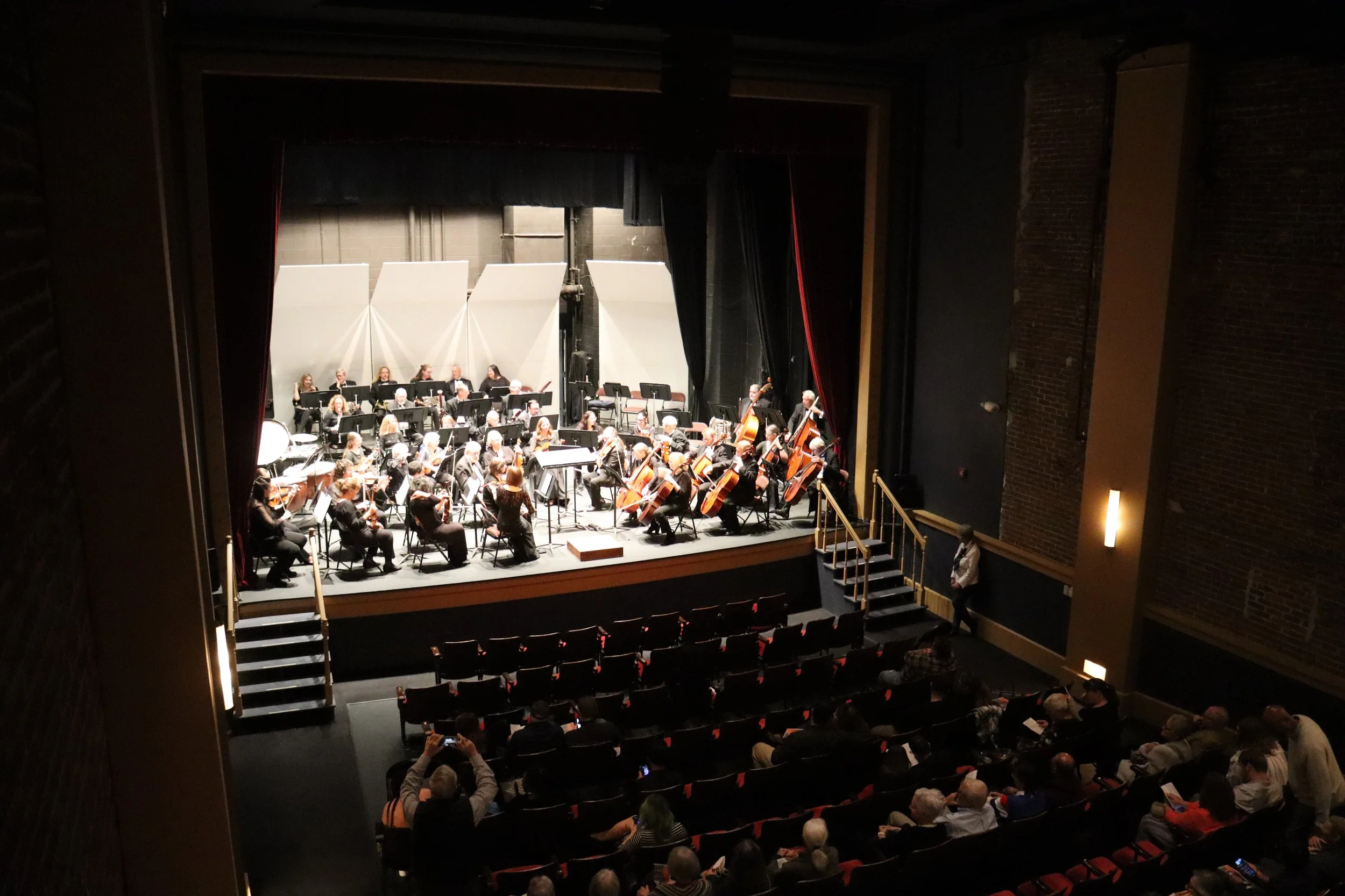 An orchestra performing on stage in a theater, with musicians playing various string instruments while the audience watches.