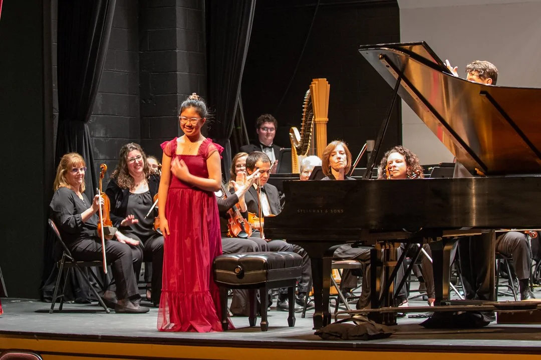 A young woman in a red dress standing on stage next to a grand piano, with an orchestra seated behind her on a black stage.