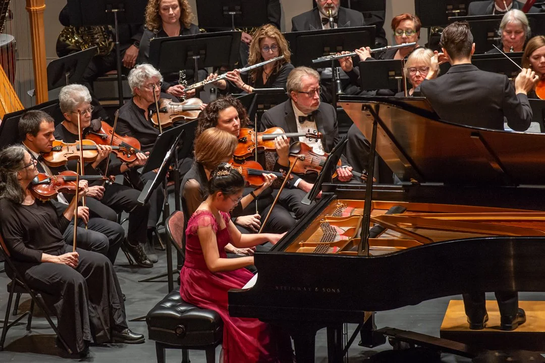 Orchestra performing on stage with a young girl playing the piano in front. The ensemble includes violinists, flutists, and other musicians, with a conductor directing.