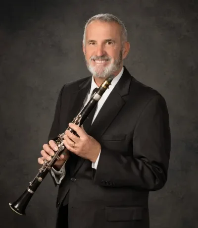 A man with a beard in a black suit holding a clarinet against a dark background.