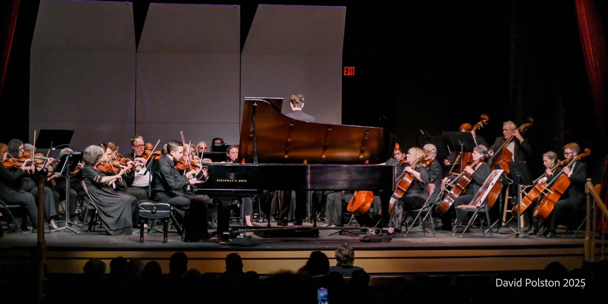 An orchestra performing on stage with a grand piano at the center, surrounded by string musicians. The stage has a dark background with an exit sign visible. Audience silhouettes are seen at the bottom.