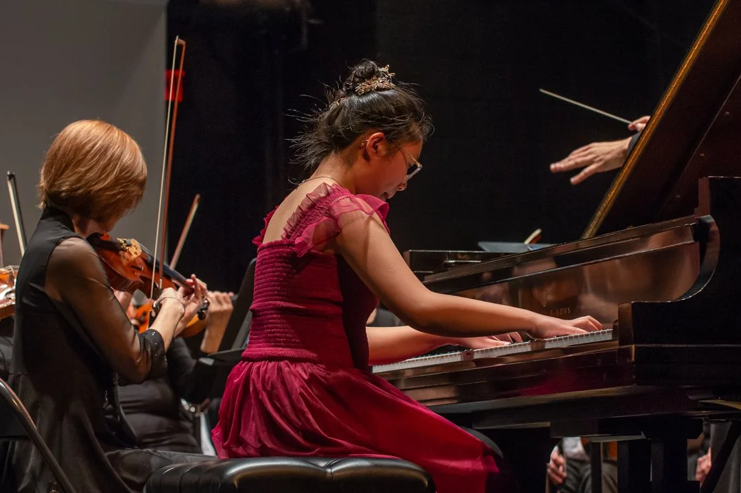 Young girl in a red dress playing the piano during a performance, with violinists in the background.