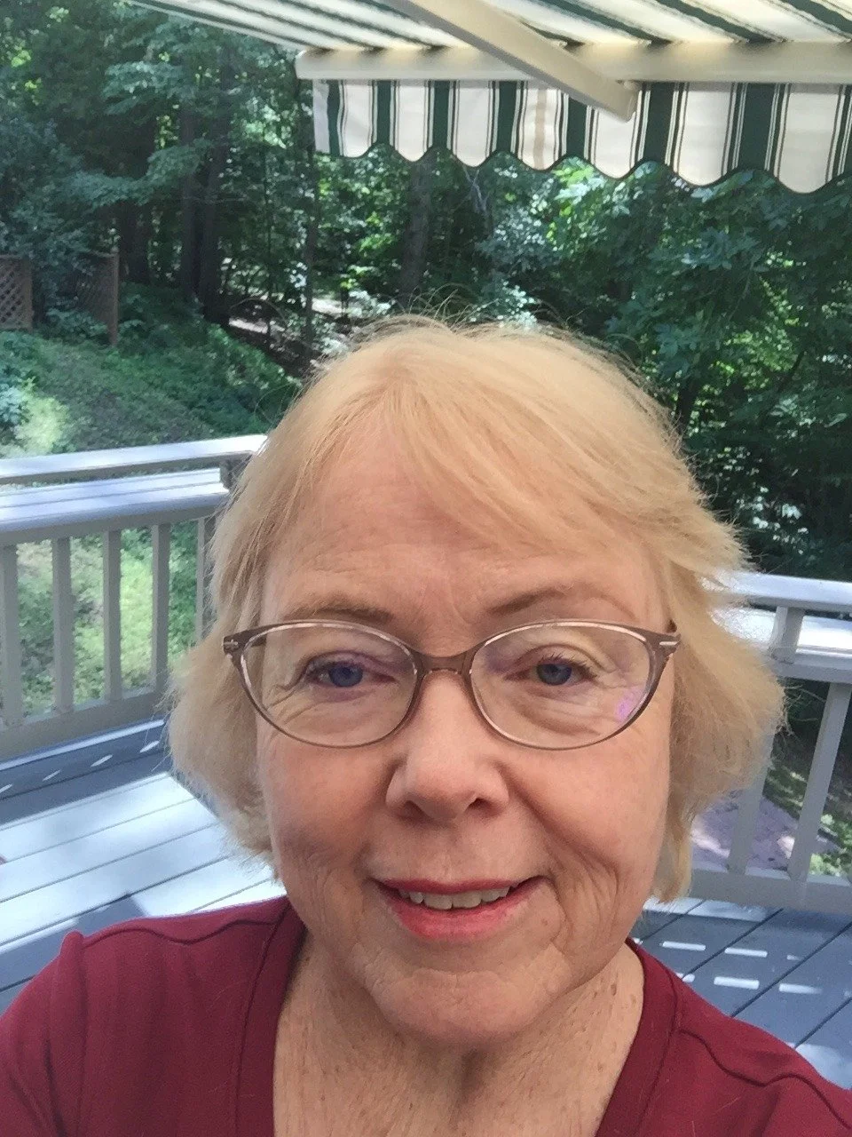 A close-up selfie of an older woman with blonde hair and glasses, smiling outdoors on a balcony with trees and foliage in the background.