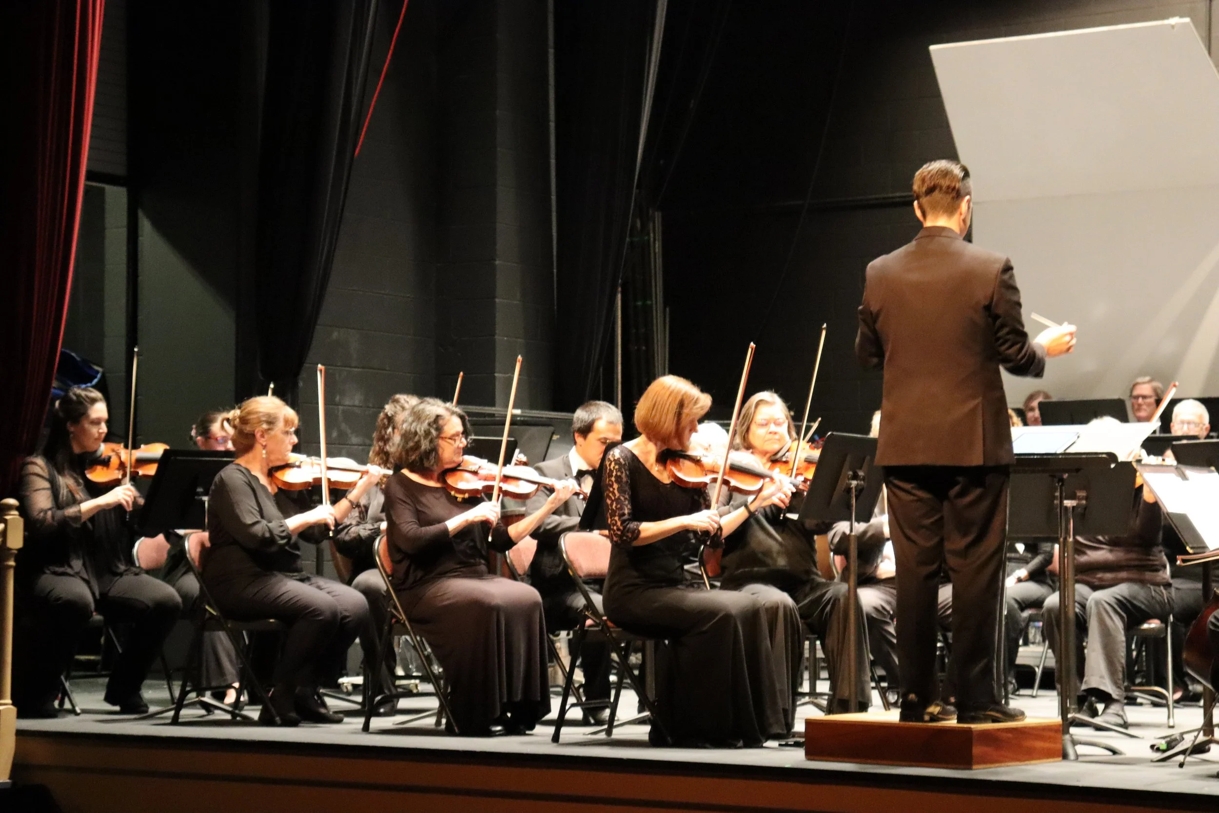 An orchestra performing on stage with a conductor in a brown suit leading them. The orchestra members are dressed in black and playing string instruments, seated in front of a dark background.