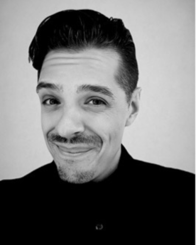 Black and white photo of a smiling man with slicked-back dark hair, a trimmed beard and mustache, wearing a dark shirt, against a plain background.