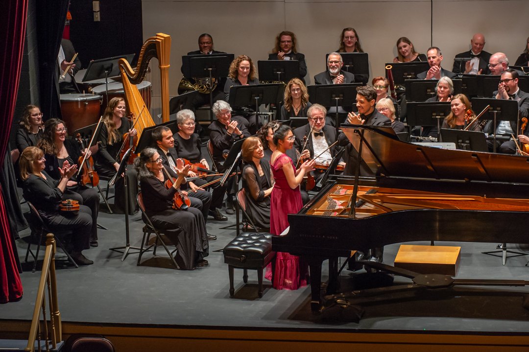 Orchestra performing on stage with a pianist, conductor, and various musicians with string, wind, and percussion instruments, all dressed in formal black attire.