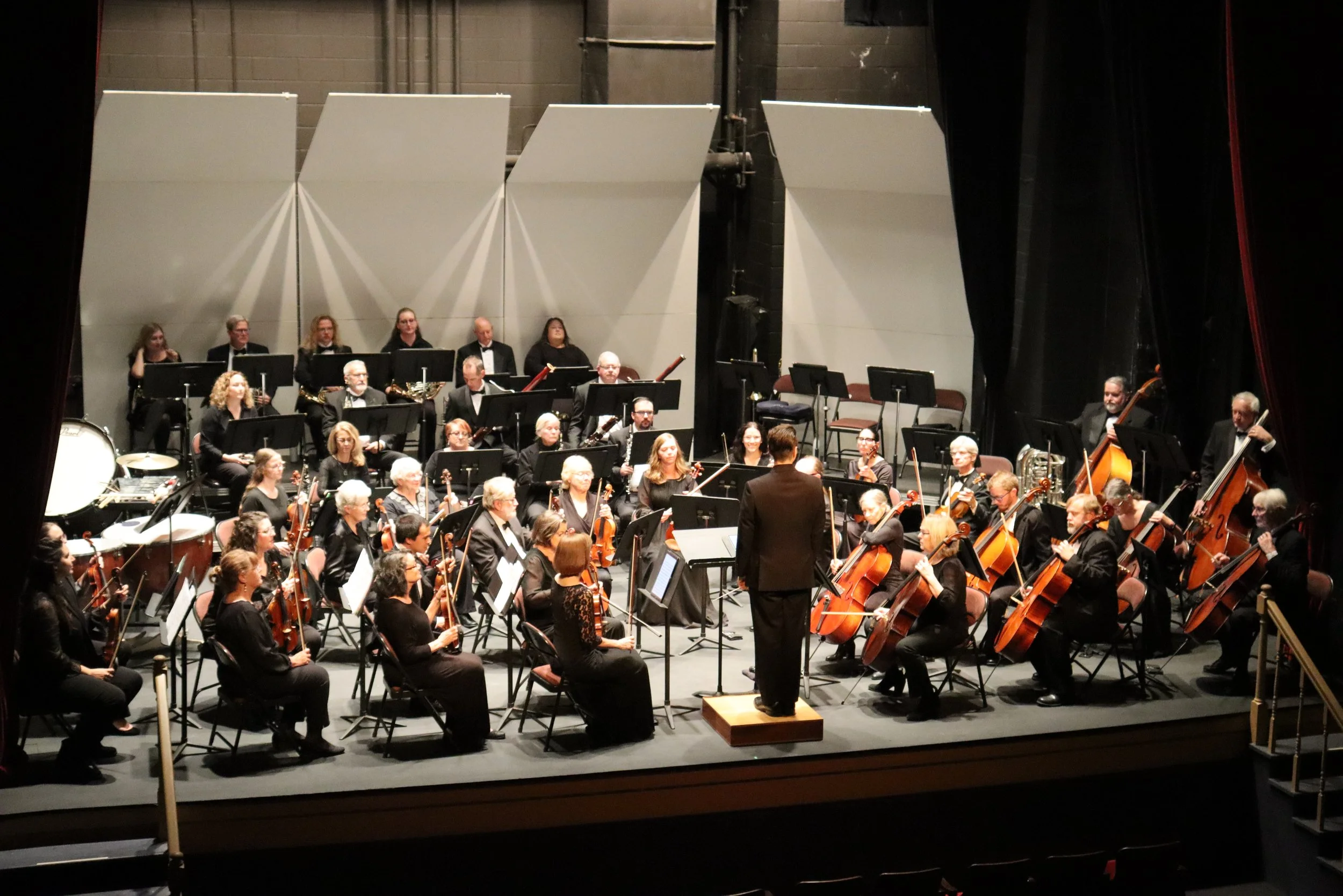 Orchestra performing on stage with conductor in front, musicians playing violins, cellos, and other instruments, black stage curtains on sides.