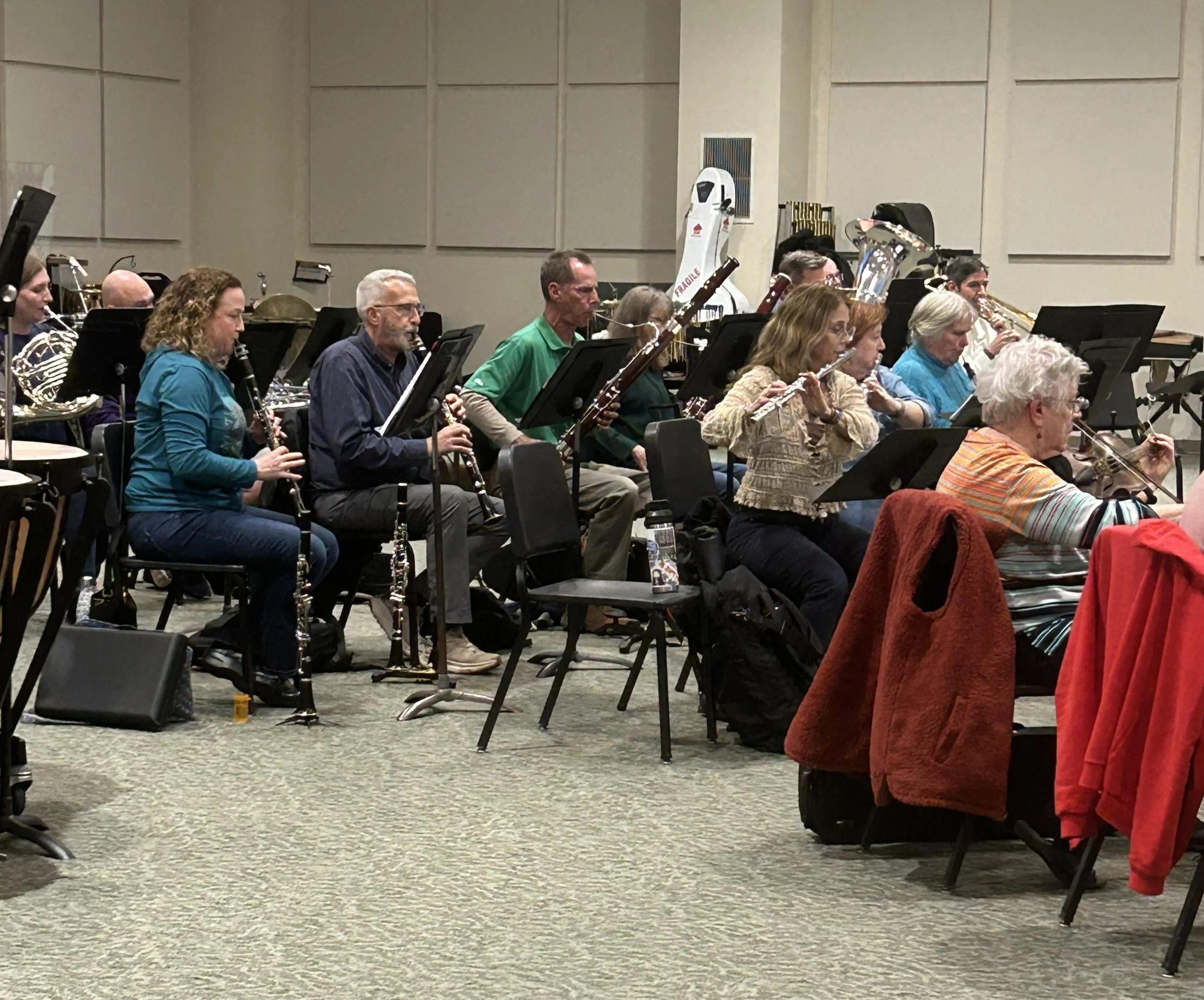 An orchestra rehearsal with musicians playing wind instruments like clarinets and flutes, sitting in rows on chairs in a large room with beige walls.