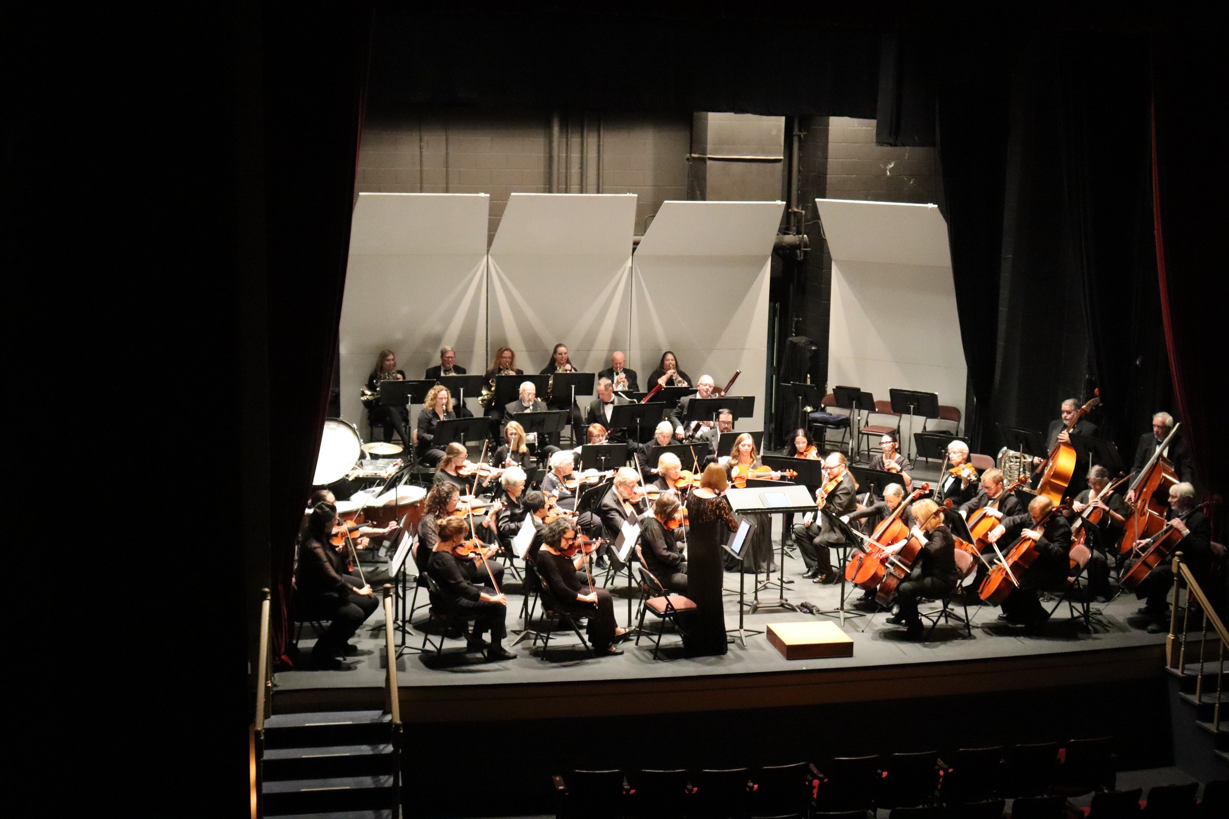 An orchestra performing on stage, viewed through an open curtain with the stage set up with musicians playing various instruments.