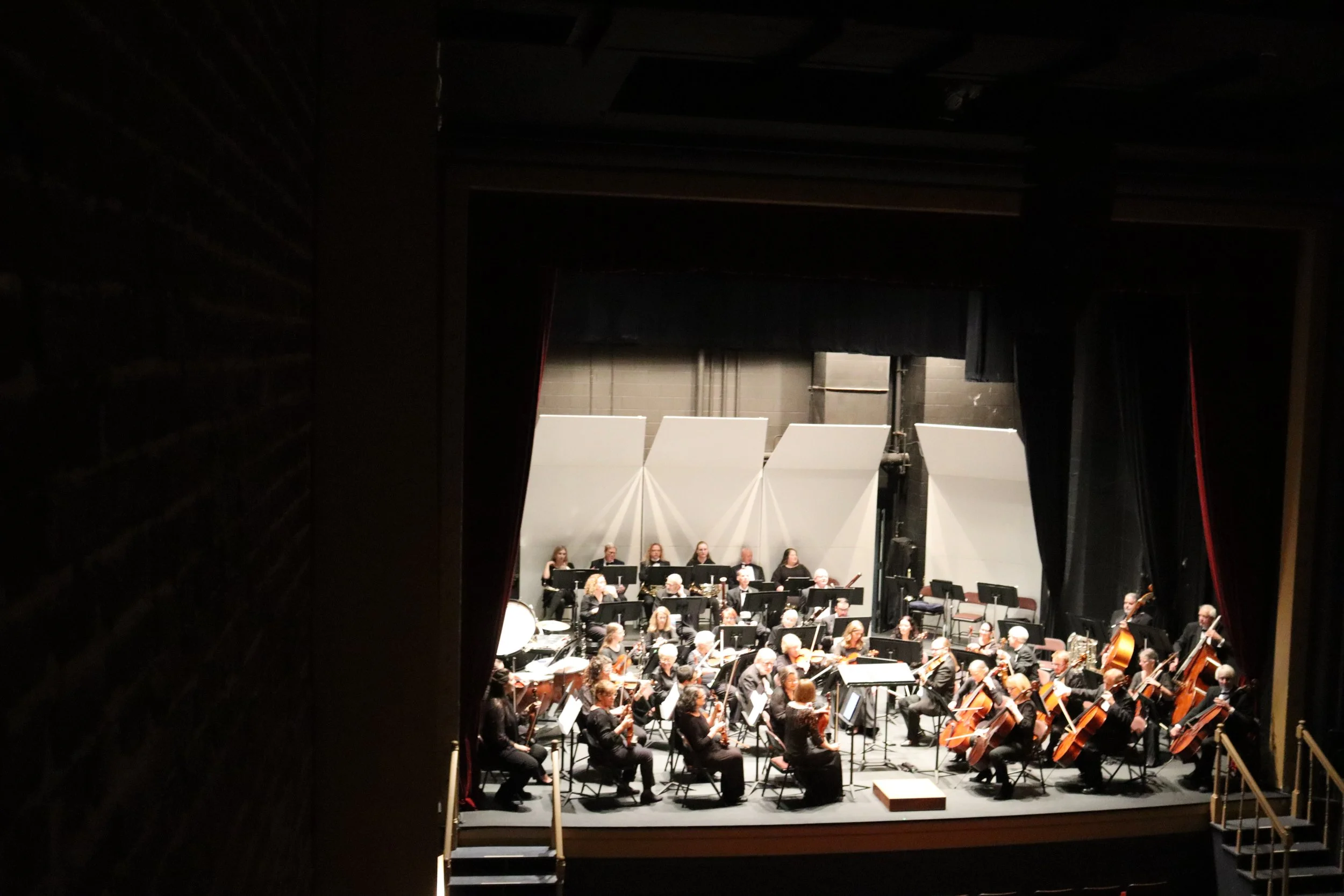 An orchestra on stage performing, with musicians playing various instruments, viewed from backstage through a partially open curtain.