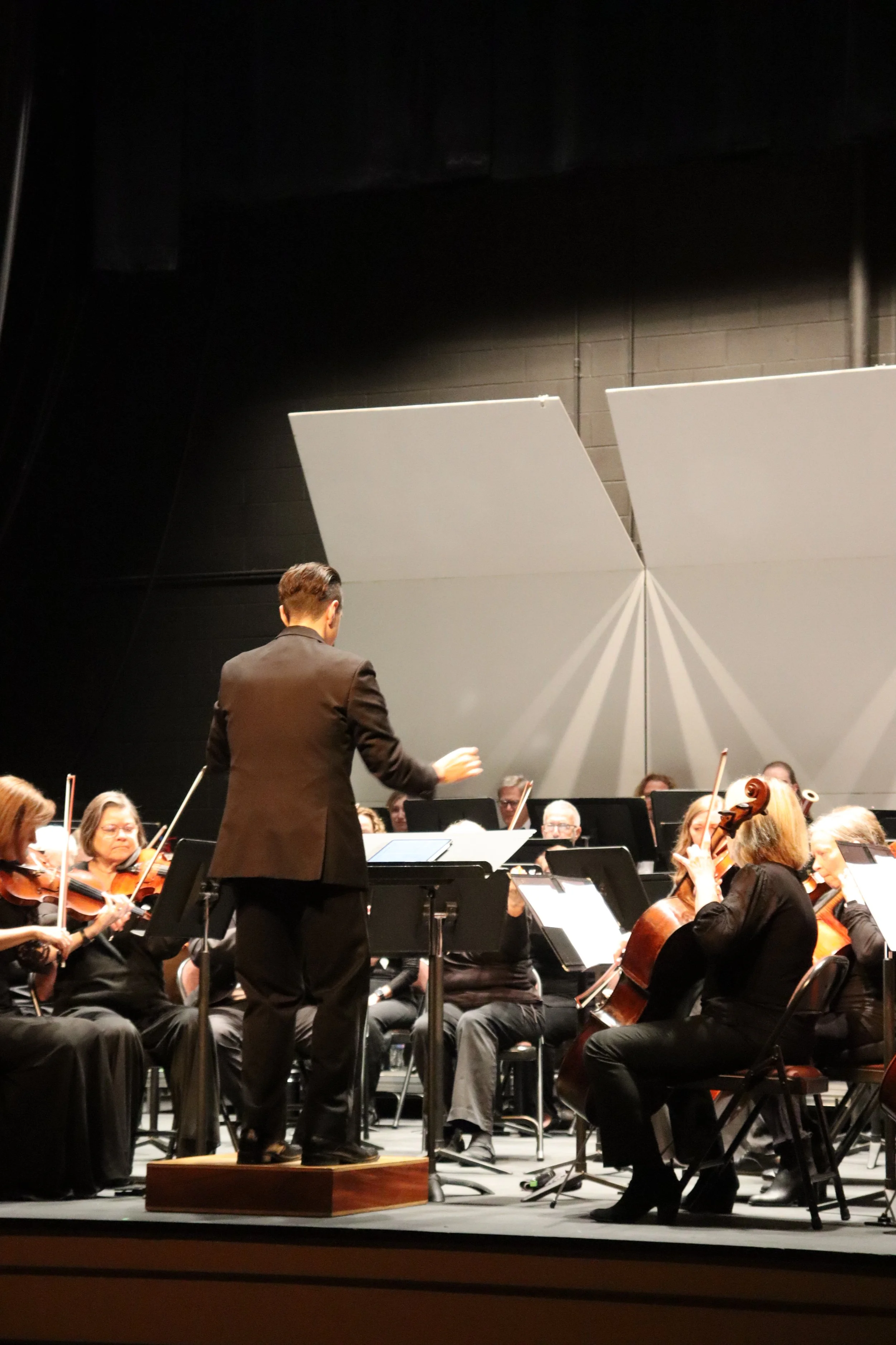 An orchestra performing on stage with a conductor standing on a wooden podium, surrounded by musicians playing instruments, including violins and cellos, in a large concert hall.