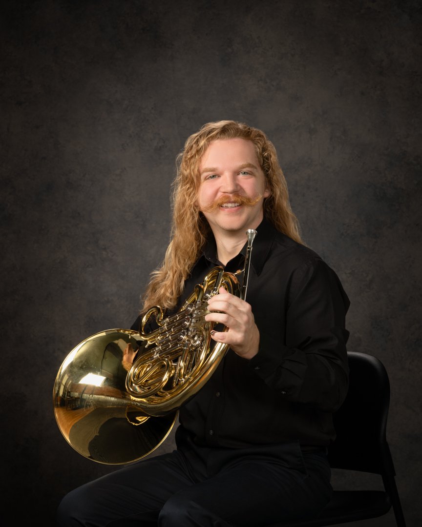 A man with long curly blonde hair and a styled mustache, wearing a black shirt, holding a French horn, sitting on a black chair, against a dark textured background.