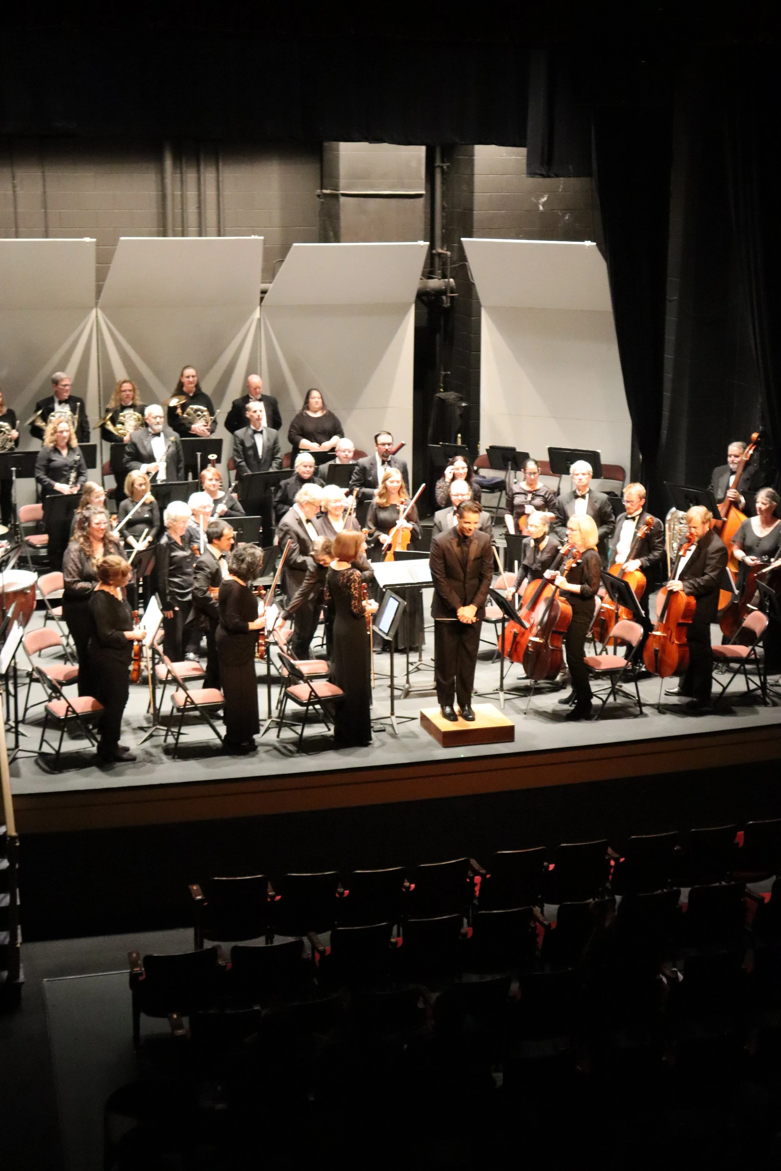 Orchestra on stage in a concert hall, with musicians seated and standing, some holding instruments, and a conductor in front.