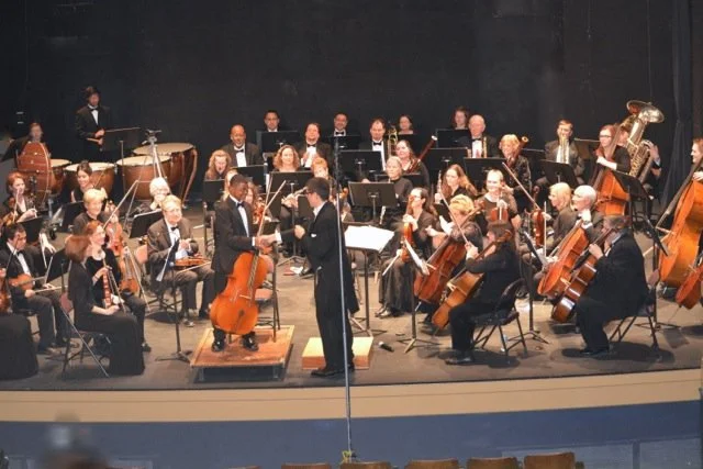 Orchestra performing on stage with conductor and various string, wind, and percussion instruments.