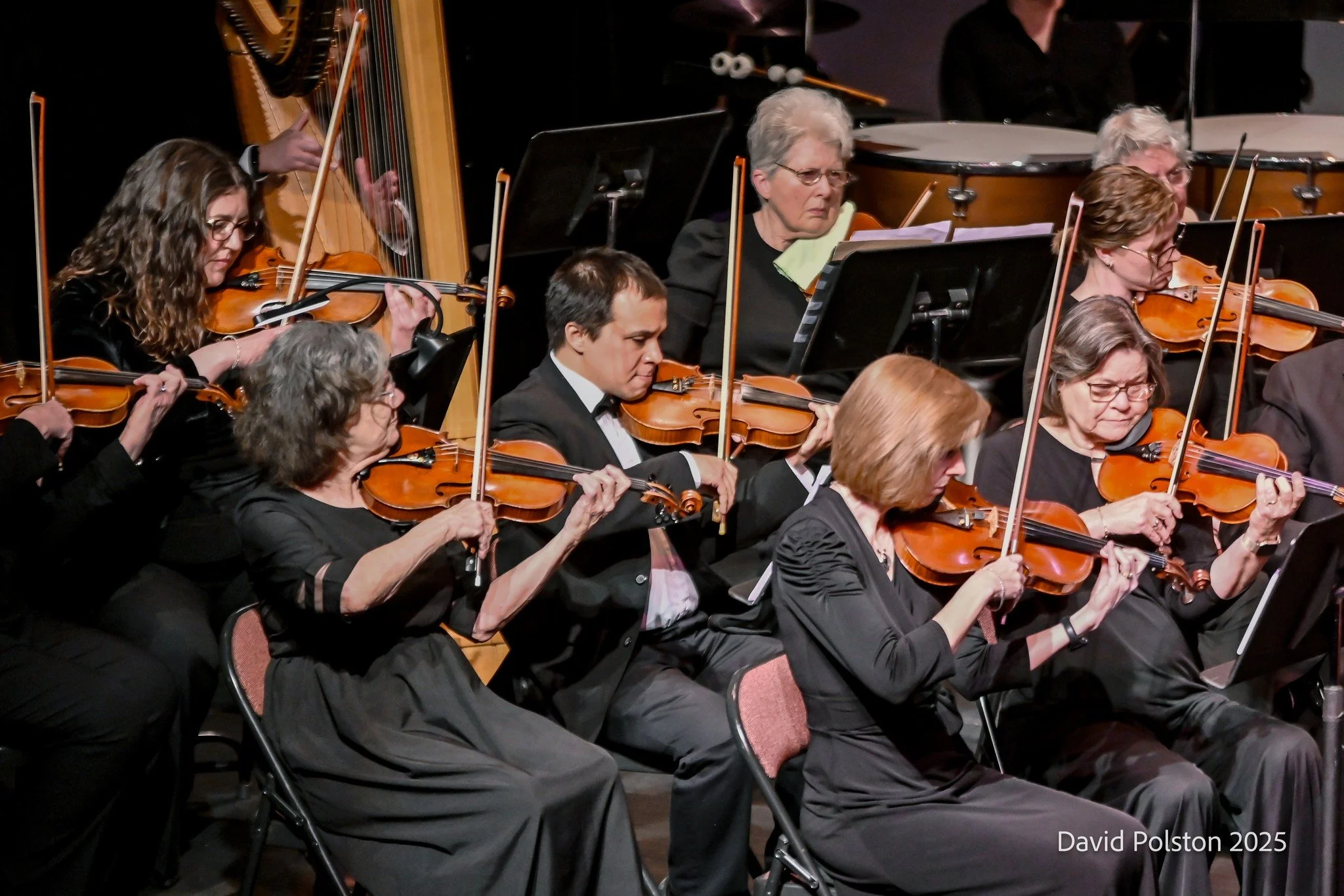 Orchestra performance with multiple musicians playing violins and other instruments on stage, some wearing glasses and formal black attire.