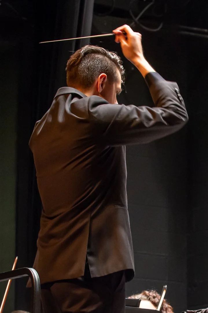 A man dressed in a black suit conducting an orchestra with a baton in his right hand, facing away from the camera.