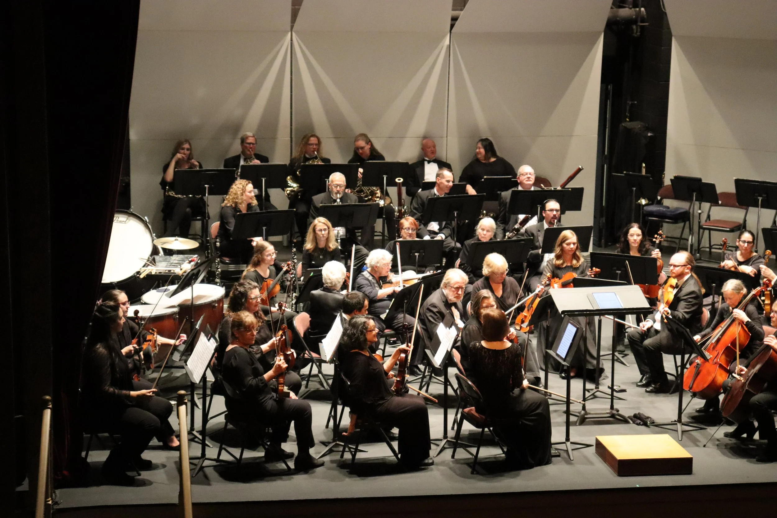 An orchestra performing on stage with musicians playing violins, cellos, and other instruments, and a group of musicians on the raised platform in the background.