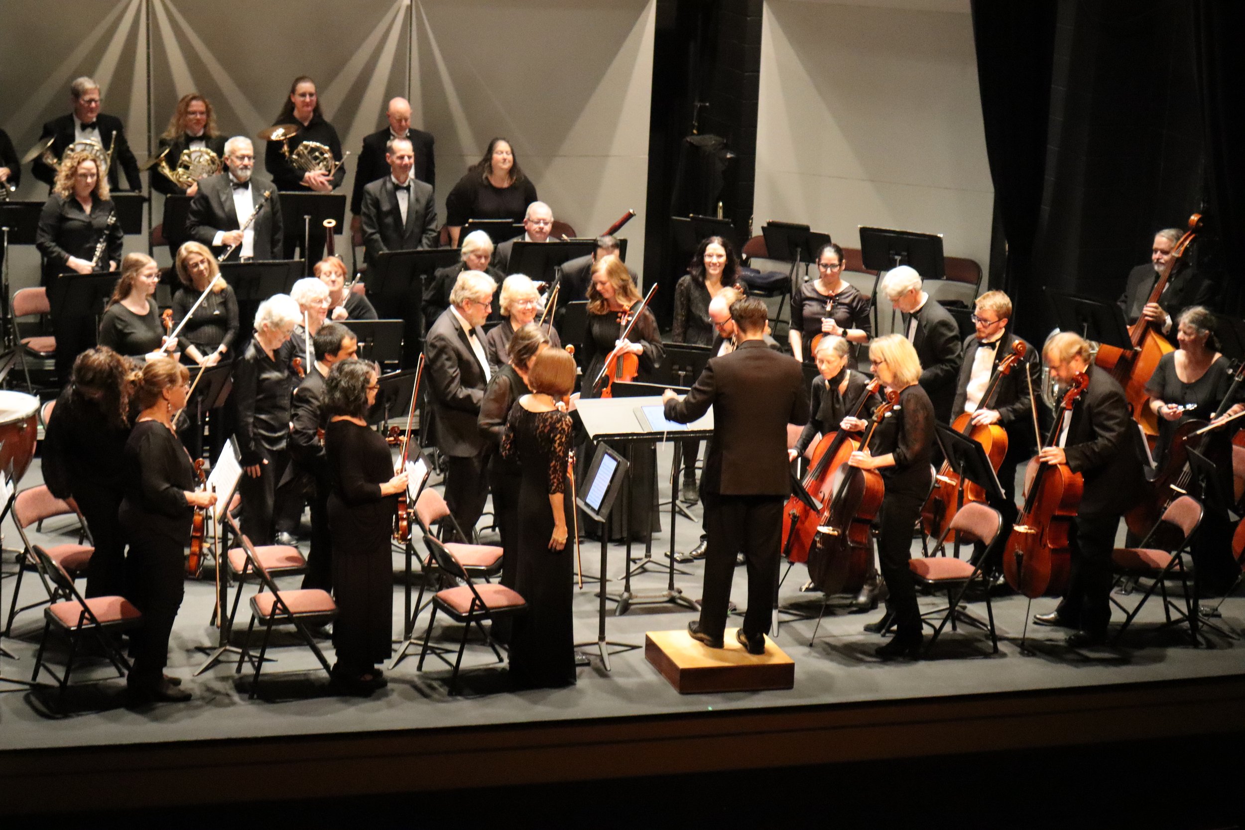 Orchestra performing on stage, with musicians holding various instruments such as violins, cellos, clarinets, and French horns. The conductor stands at the front, leading the musicians. The performers are dressed in formal black attire, and some are 