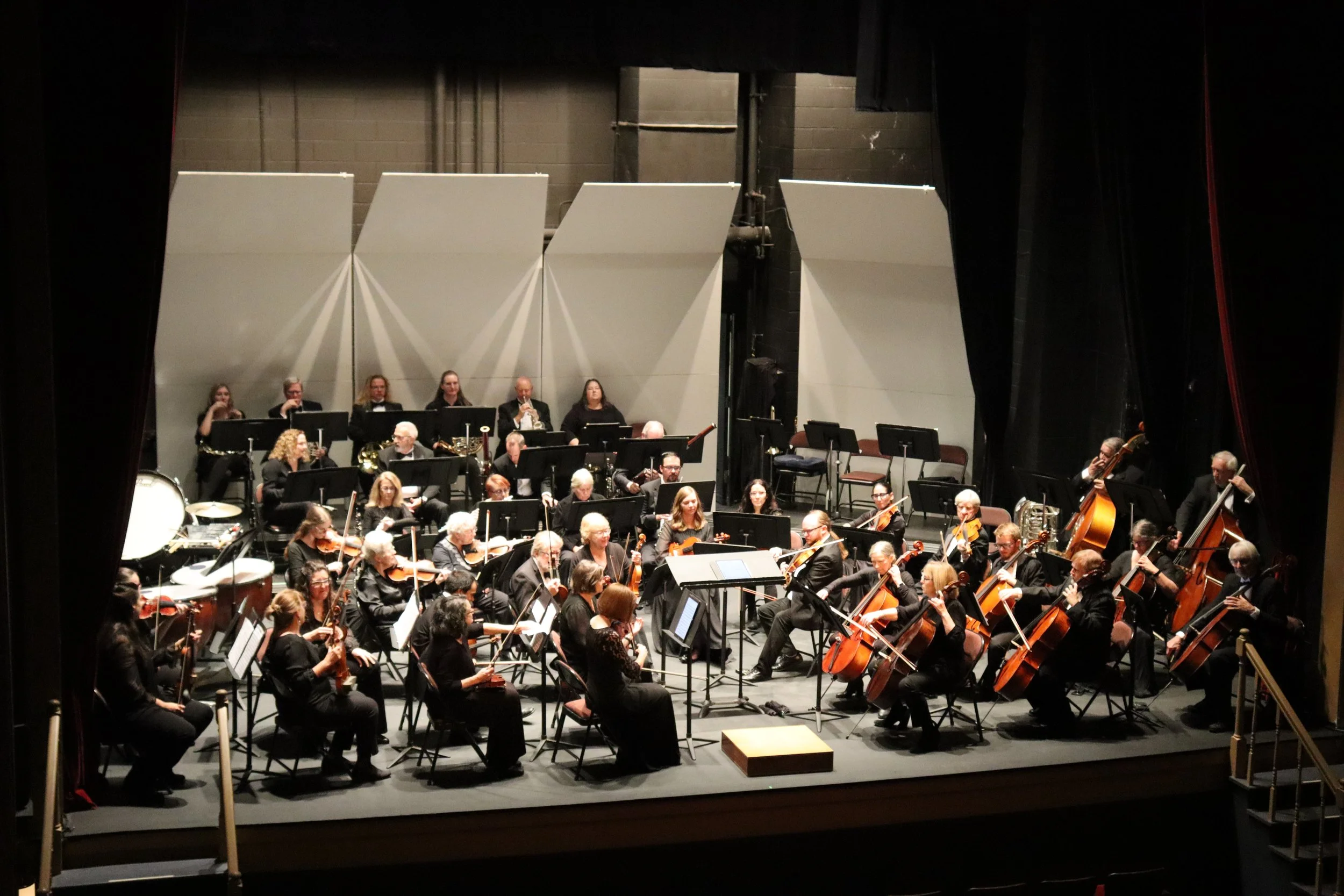An orchestra performing on stage with musicians playing various instruments, viewed through a partially open stage curtain.