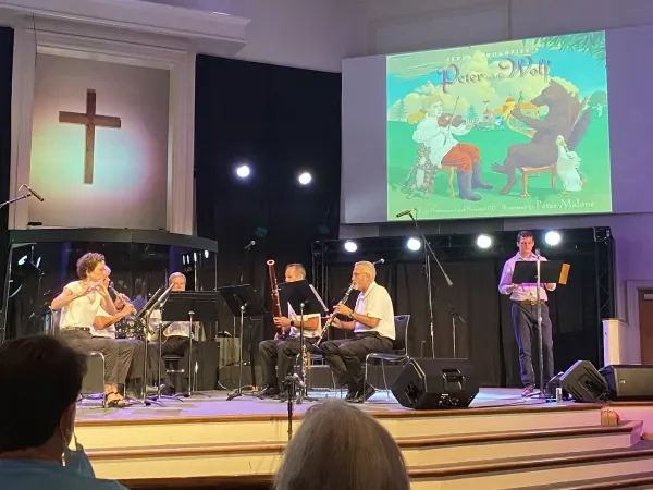 A musical performance on stage in a church, with four musicians playing instruments and a man reading from a music stand. A large screen behind them displays a colorful illustration from a storybook titled 'Peter Wool,' featuring a young girl and a bear. A cross is mounted on the wall to the left of the stage.